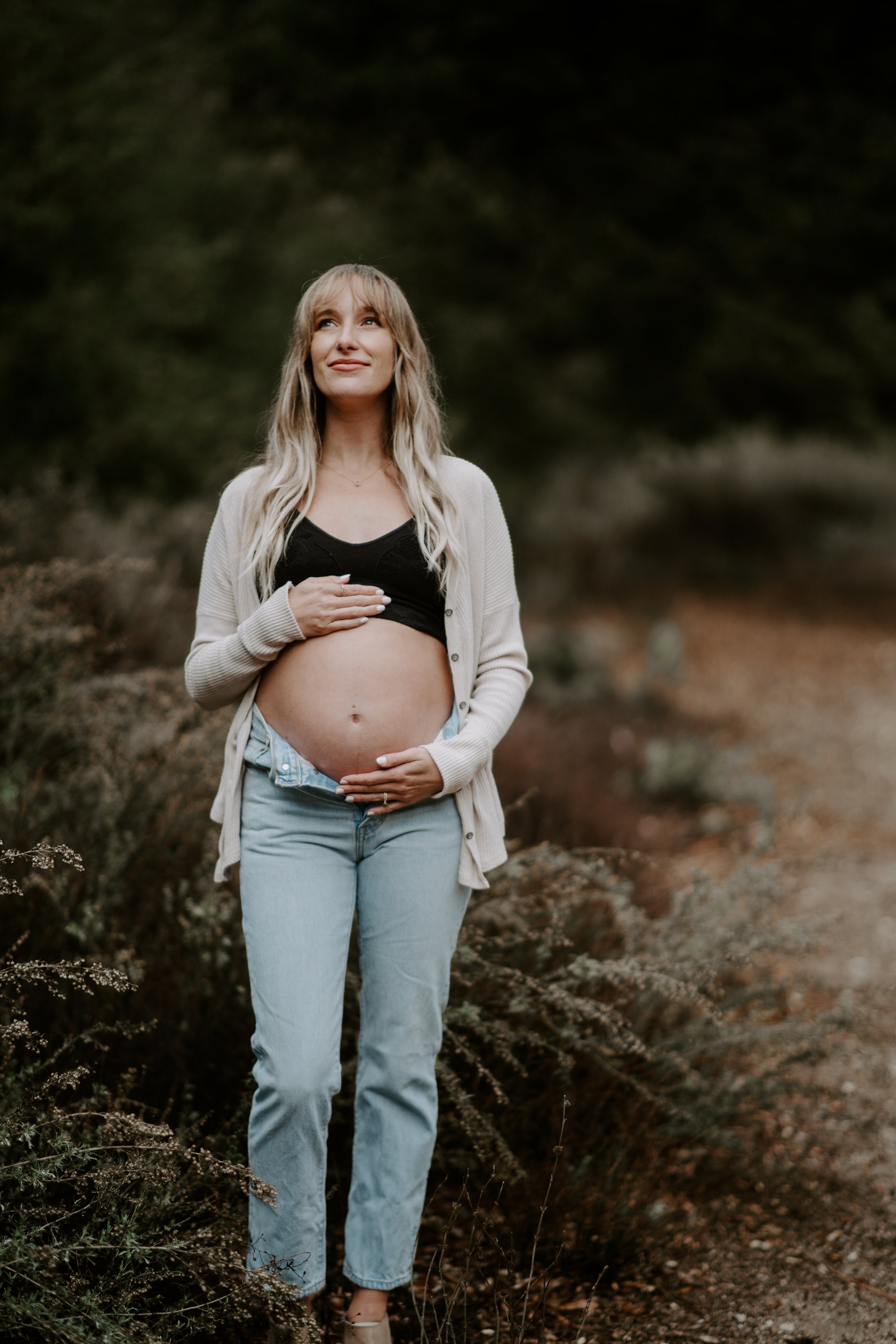 A pregnant woman standing outdoors on a natural trail, wearing a black top, a beige cardigan, and light blue jeans, gently holding her belly with a serene expression.