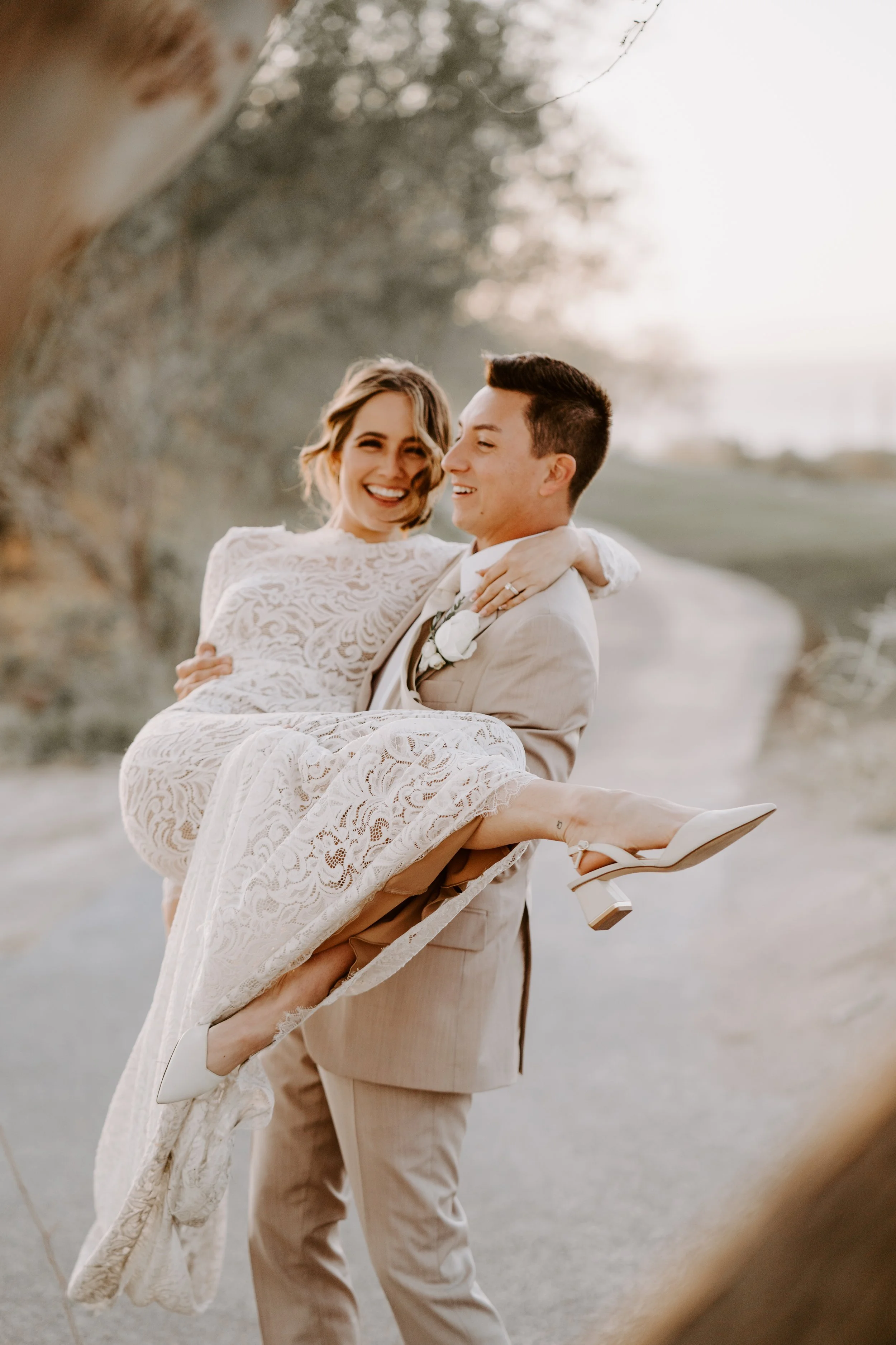 A groom holding a smiling bride in a white lace wedding dress outdoors during sunset.