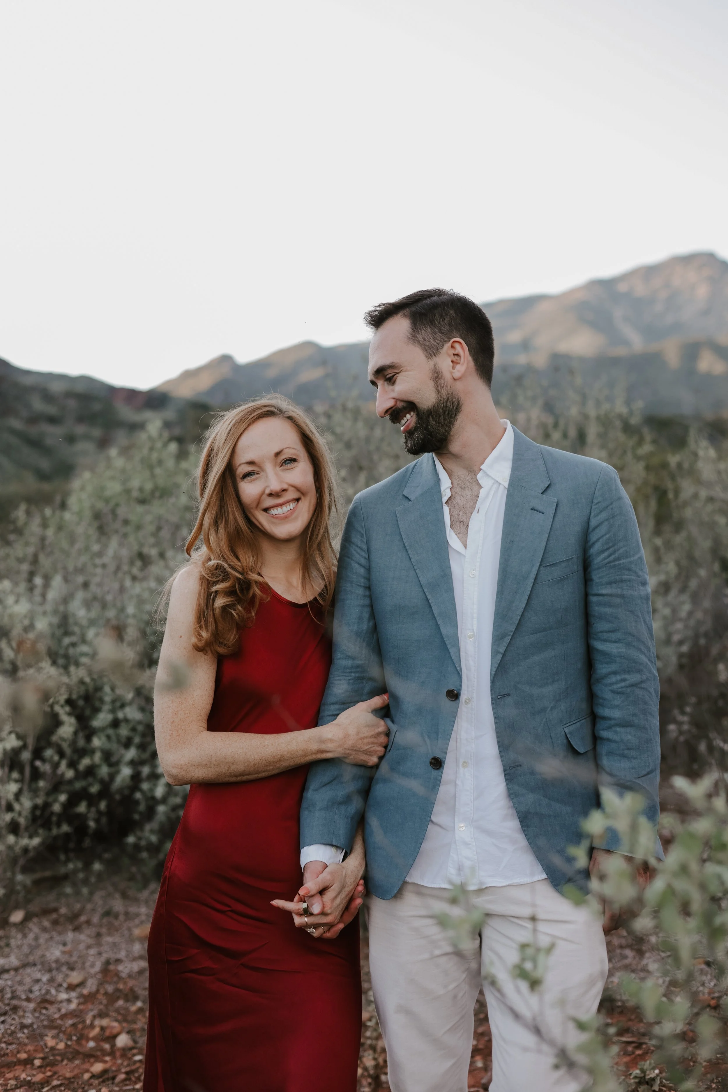 A smiling woman in a red dress holding hands with a man in a blue blazer and white shirt, standing outdoors with mountains and greenery in the background.