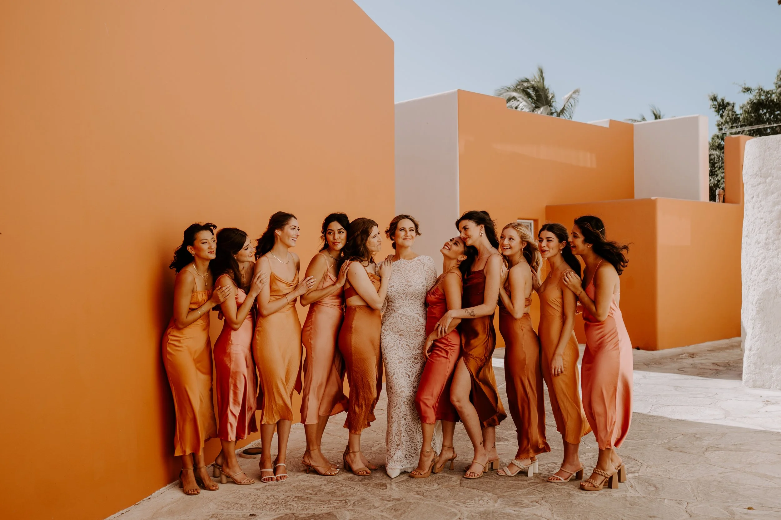 A group of women in colorful dresses gathered outdoors, posing together with smiles against an orange wall on a sunny day.