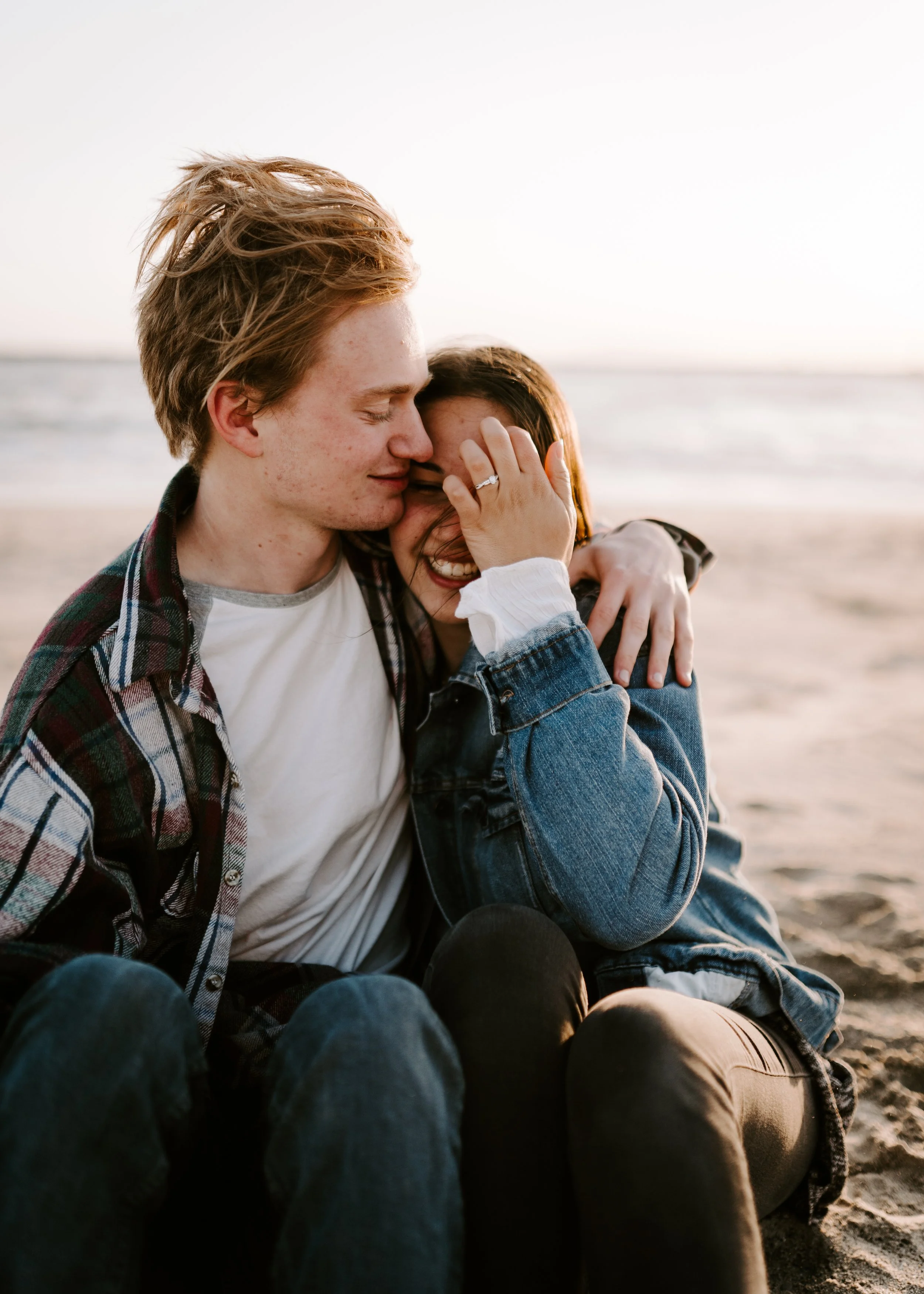 A smiling couple sitting on a beach, hugging and enjoying each other during sunset.