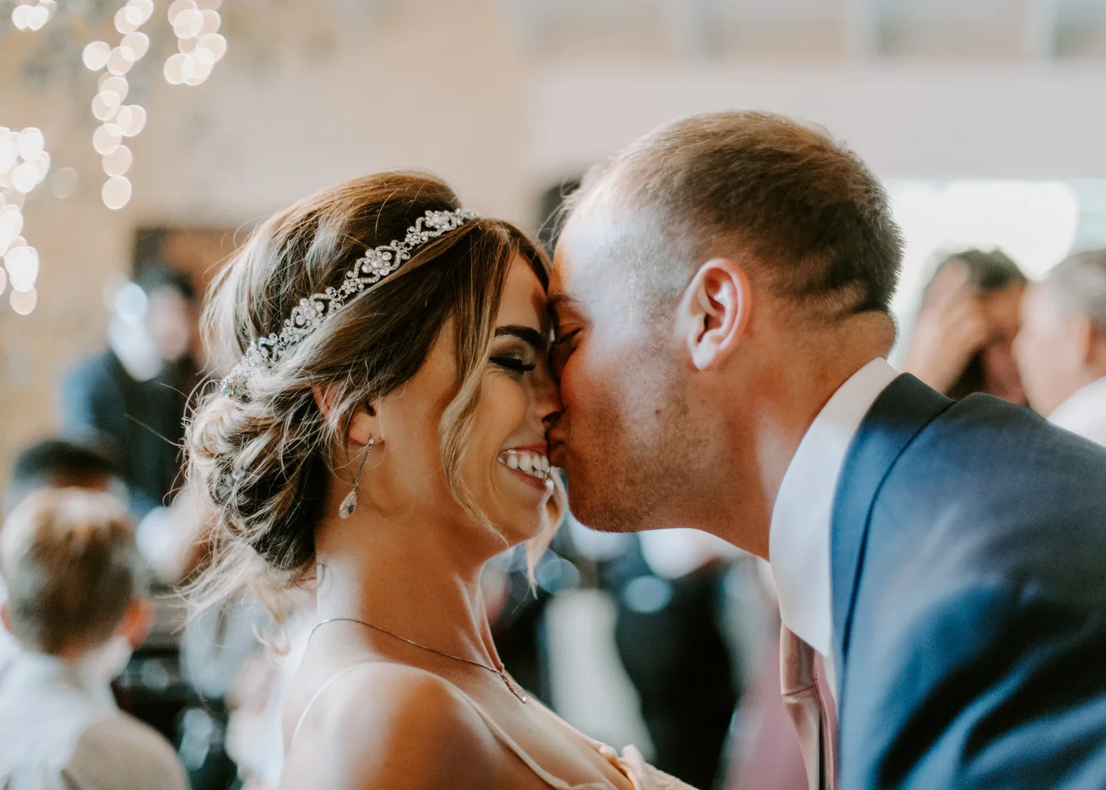 A bride and groom sharing a kiss during their wedding reception, with blurred guests in the background.