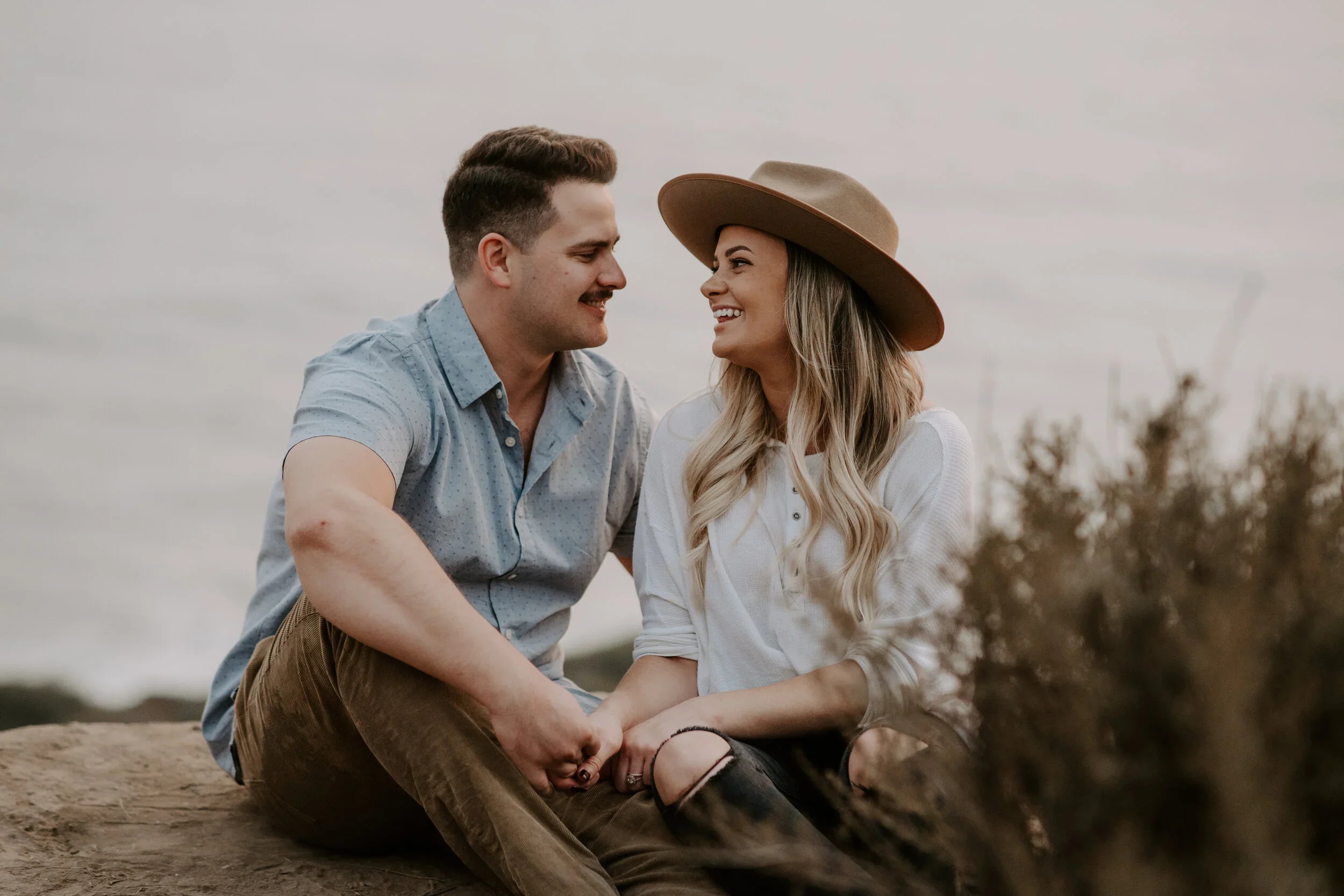A young couple sitting on a rock outdoors, smiling at each other. The man is wearing a short-sleeved, light blue shirt, and the woman is wearing a white shirt and a wide-brimmed hat. They are holding hands, and the background is blurred with cloudy sky and some bushes.