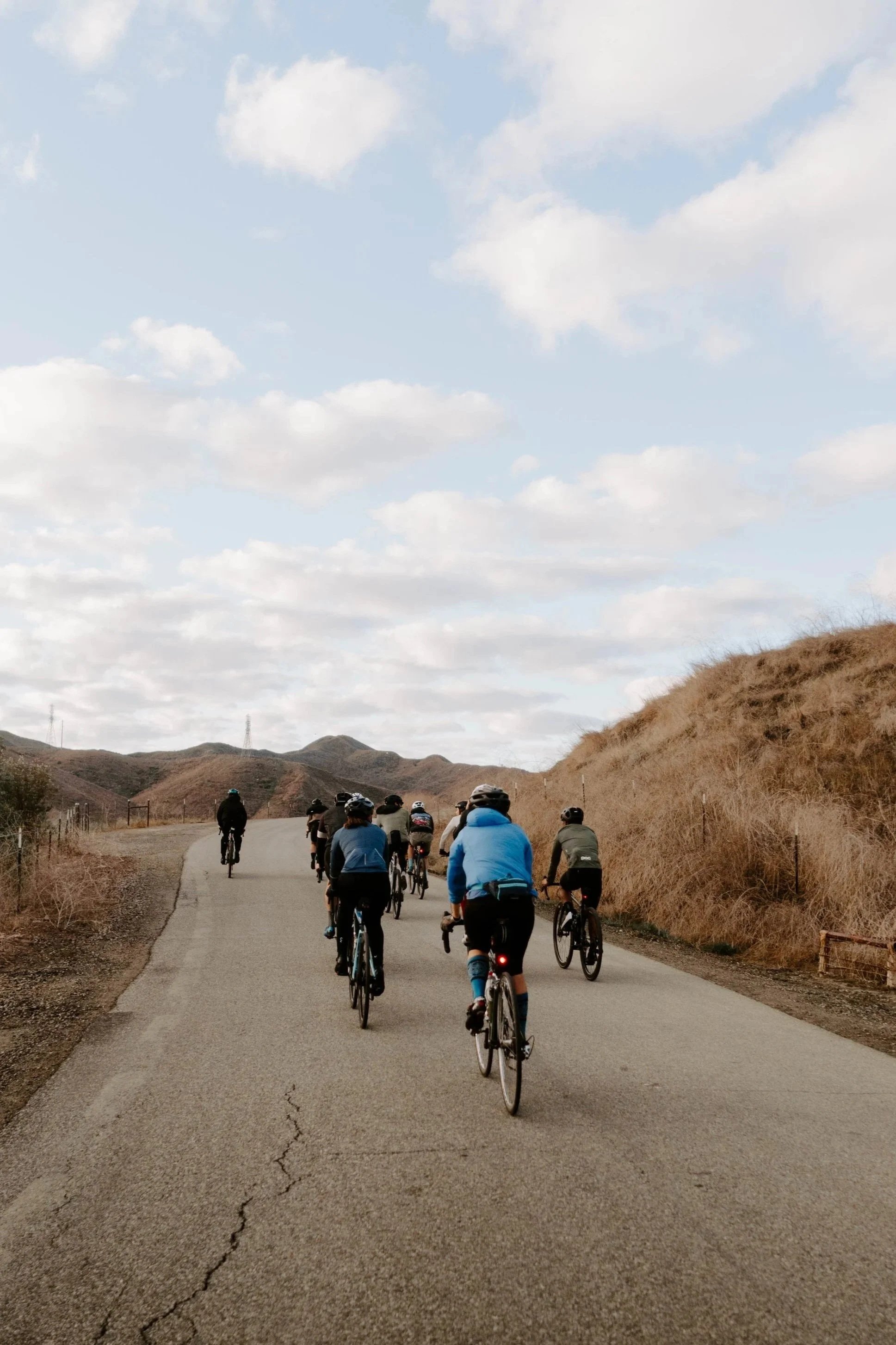 Group of cyclists riding on a paved road through hilly landscape under partly cloudy sky.