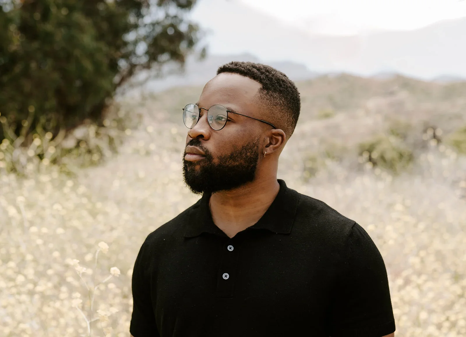 A man with glasses and a beard wearing a black polo shirt, standing outdoors in a natural setting with trees and hills in the background.