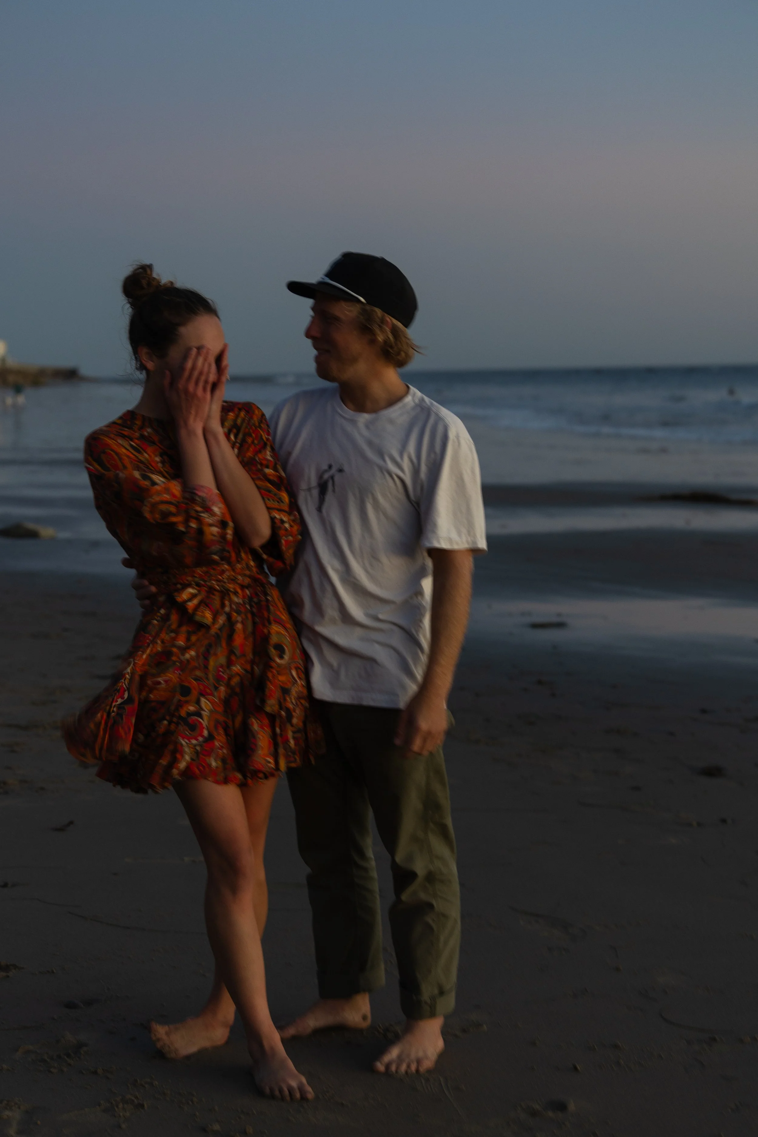 A young woman with her hands over her face smiling as a young man in a cap and casual clothes looks at her while standing barefoot on the beach during sunset.