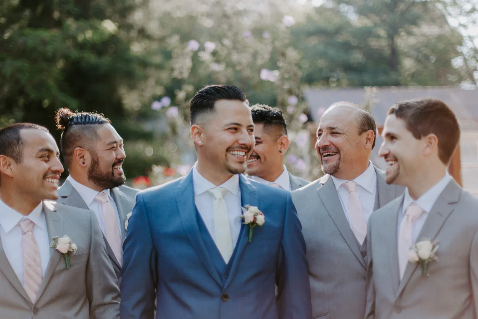 A group of six men in suits smiling and laughing outdoors during a wedding celebration, with a groom in a blue suit and five groomsmen in light gray suits and pink ties, standing in front of a tree with purple flowers.