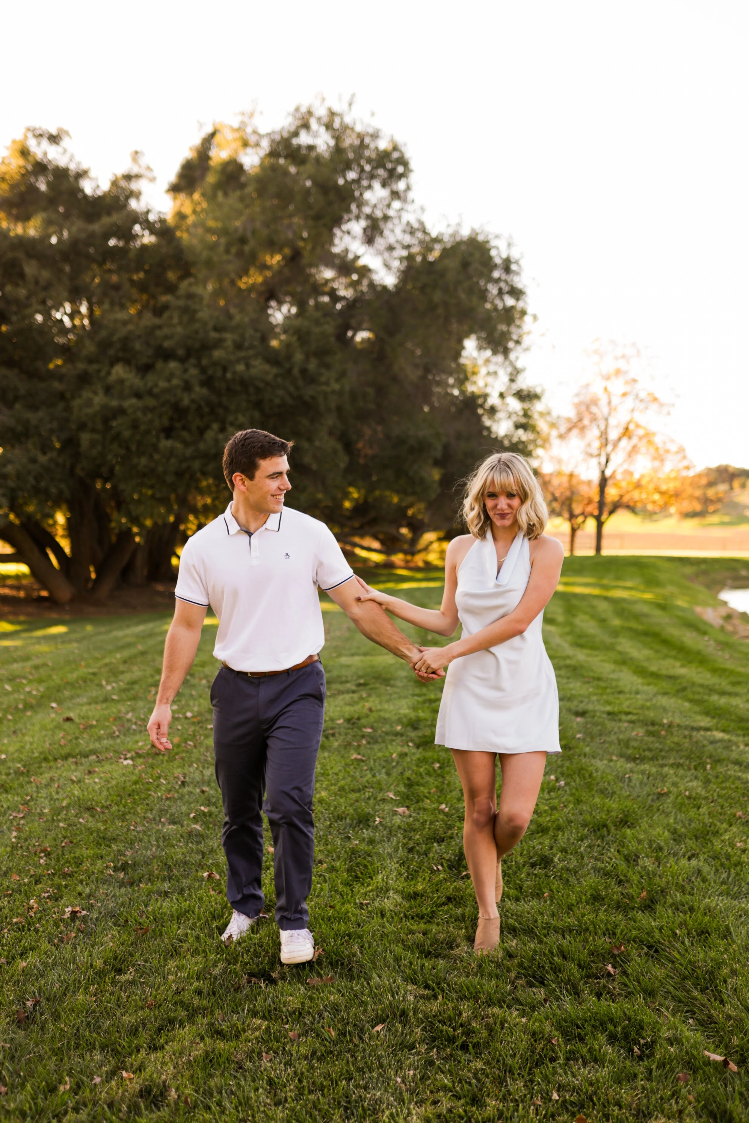 A young couple holding hands, walking on a grassy field at sunset, with trees in the background.