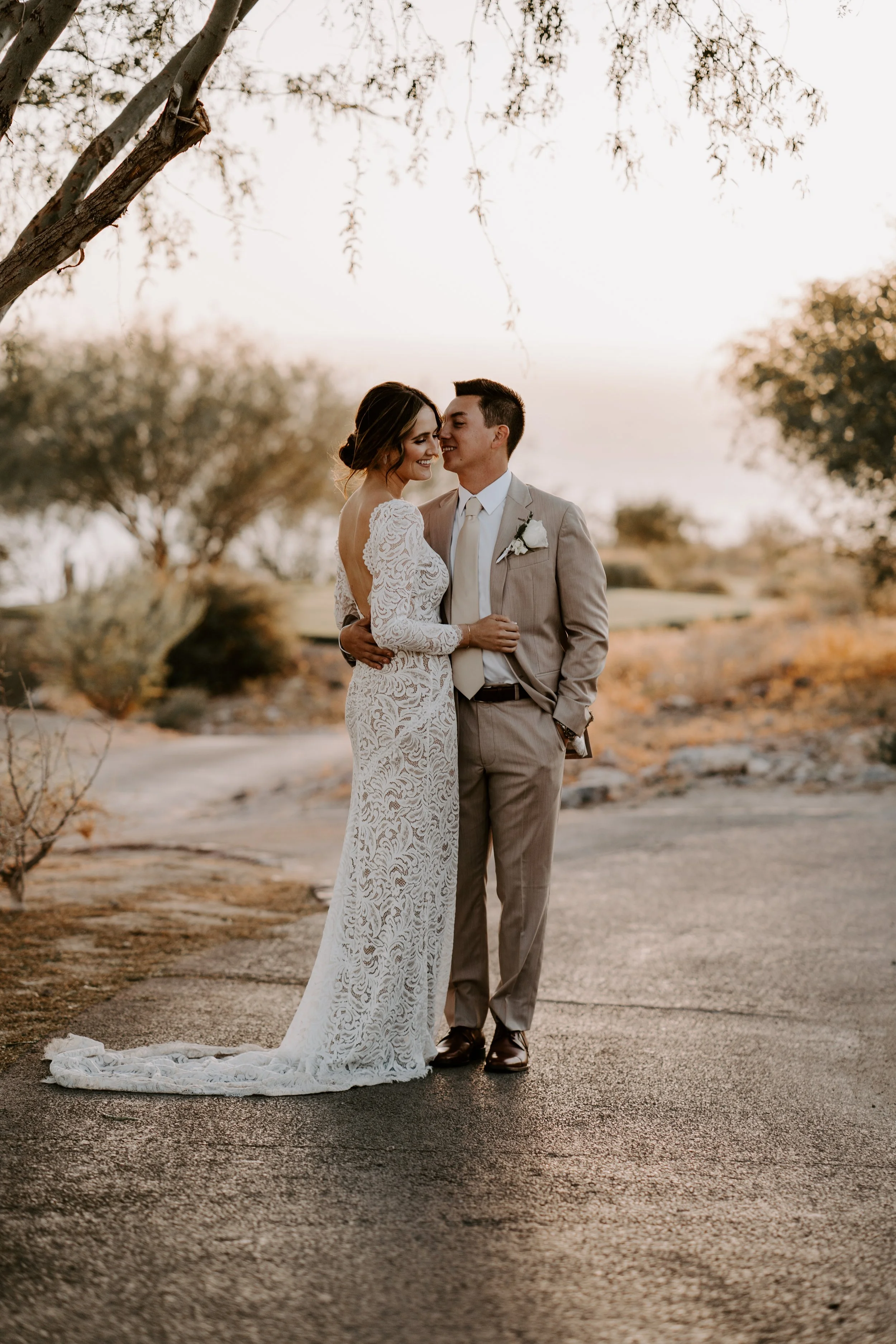 A bride and groom stand close together outdoors during sunset, smiling at each other, with a tree overhead and desert landscape in the background.