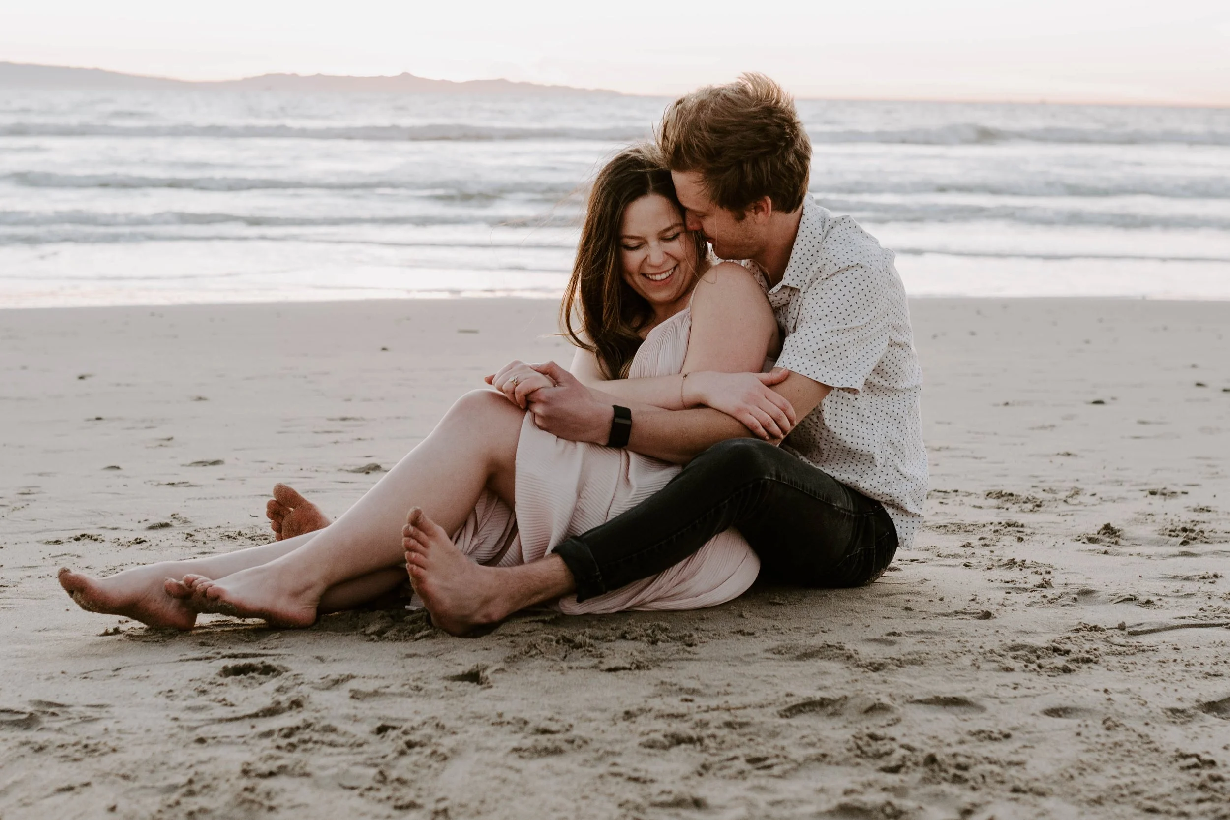 A happy couple sitting on the beach at sunset, embracing and smiling at each other.