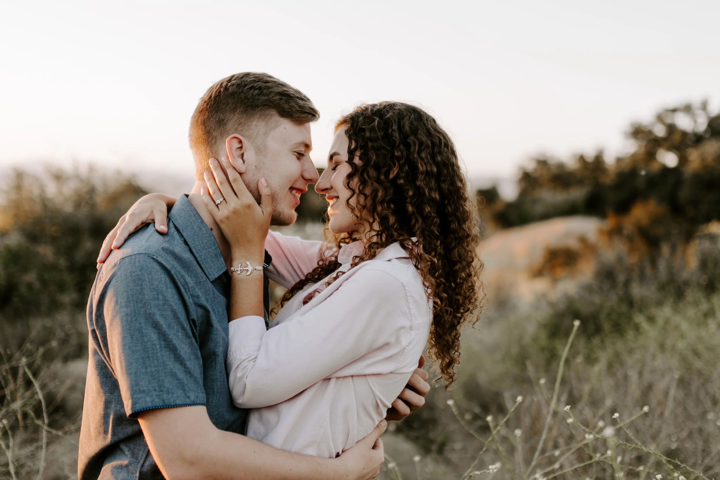 A young couple embraces outdoors during sunset, with their foreheads touching, smiling, and holding each other tenderly.