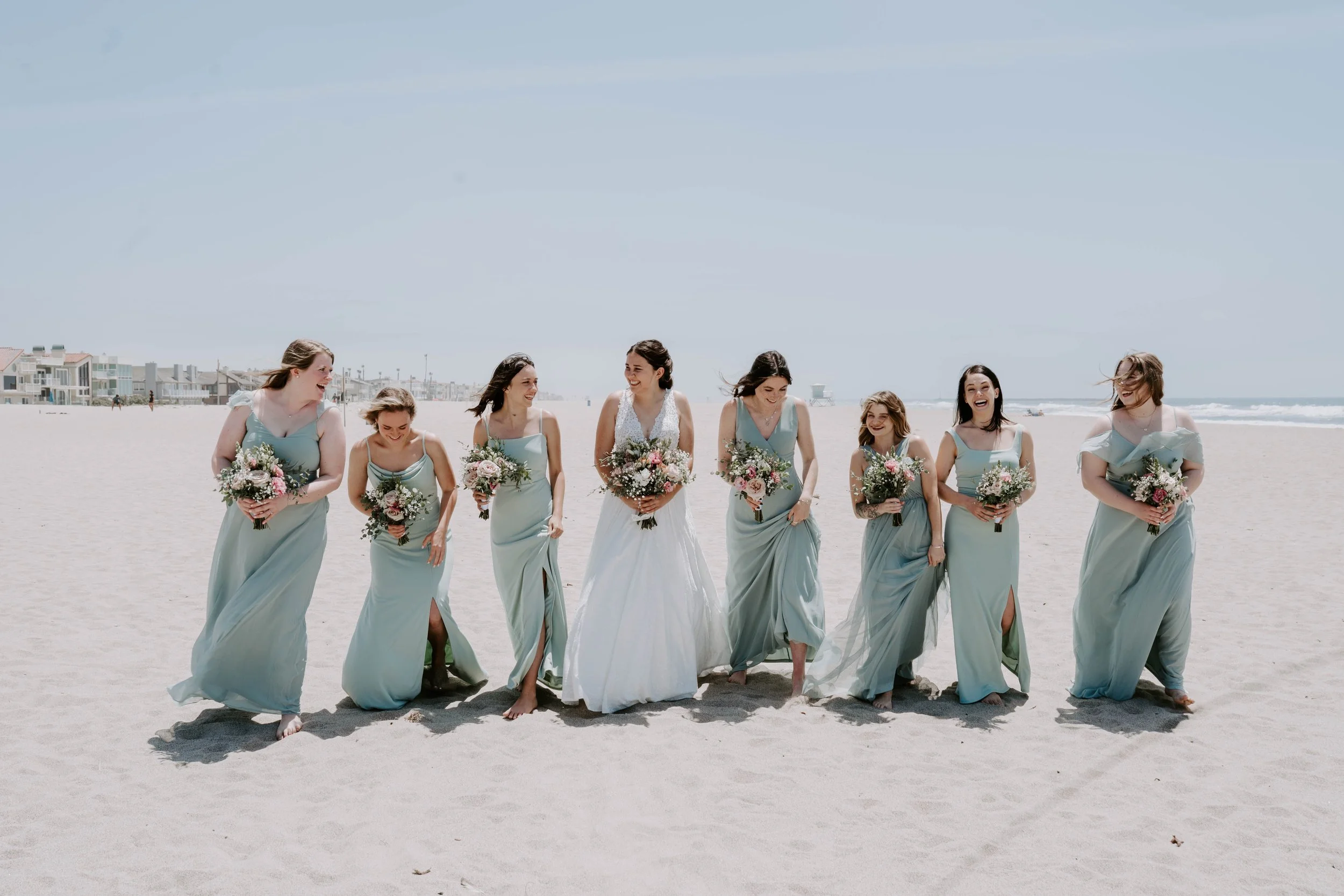 Bride and seven bridesmaids walking barefoot on the beach, holding bouquets of flowers, with the ocean and beach houses in the background.