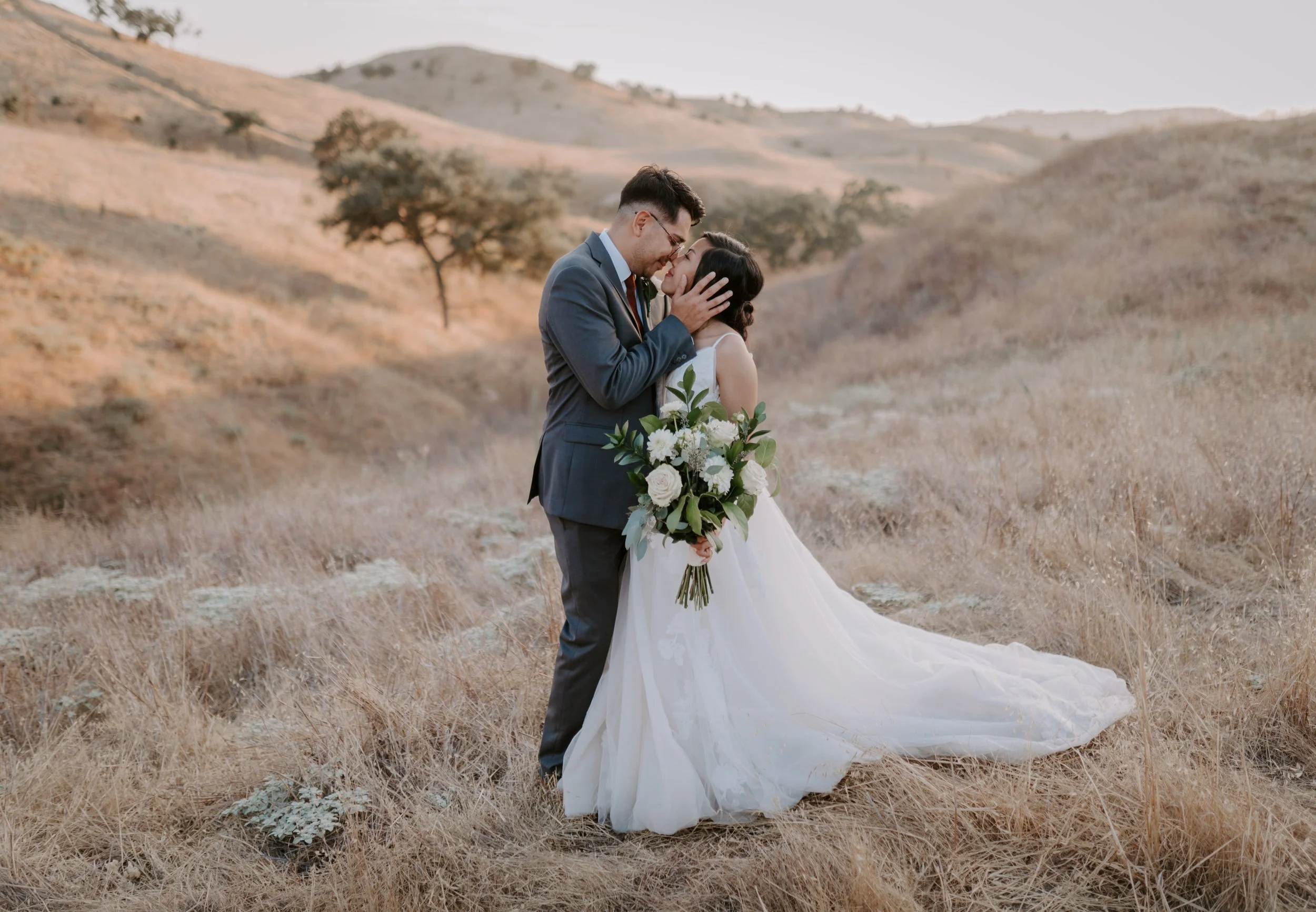 A newlywed couple stands in a dry, grassy field with rolling hills and sparse trees in the background, sharing an intimate moment with their foreheads touching. The bride is holding a bouquet of white flowers and greenery, and the groom is gently hol
