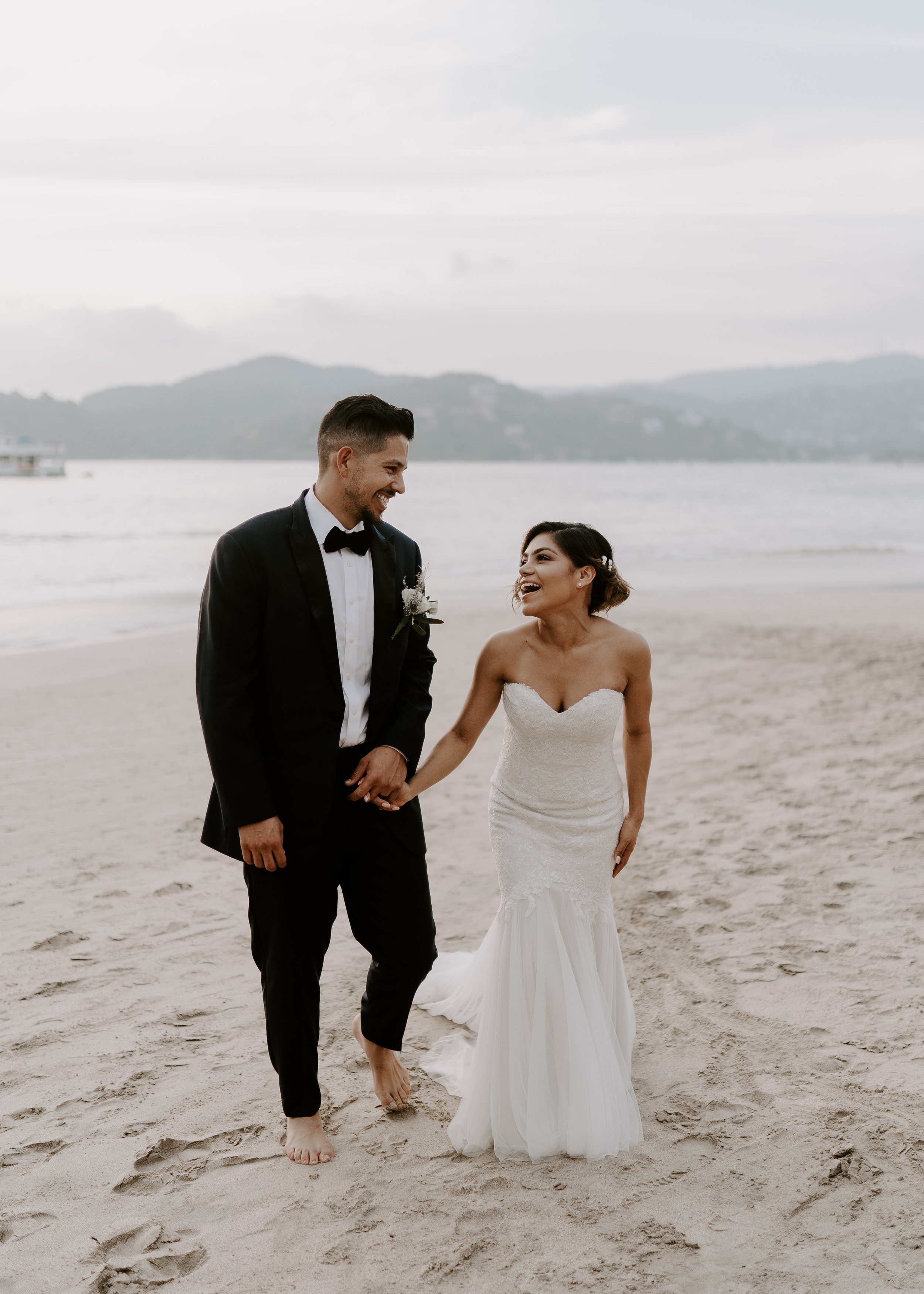 Happy couple in wedding attire holding hands and walking barefoot on the beach, smiling at each other, with ocean and mountains in the background.