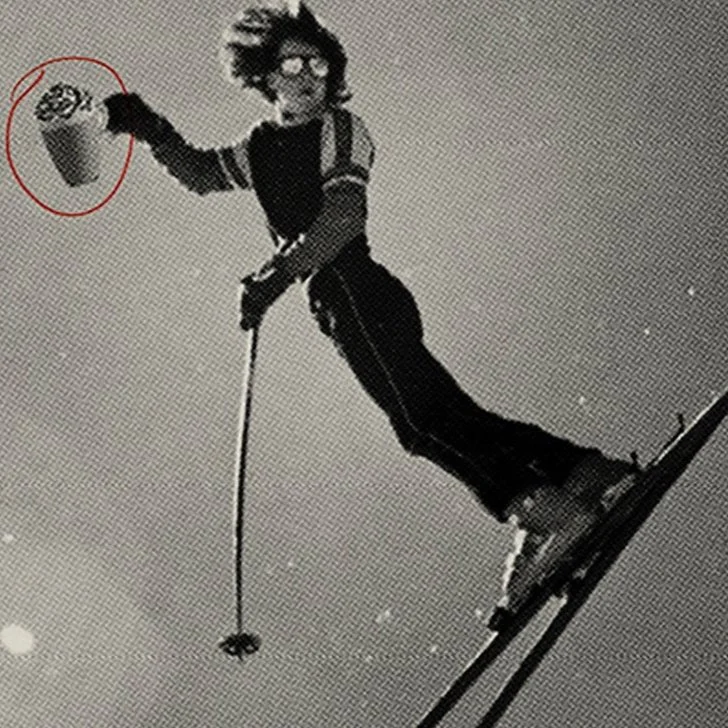 A person dressed as a cartoon character, standing on skis in midair, holding a bucket in one hand.
