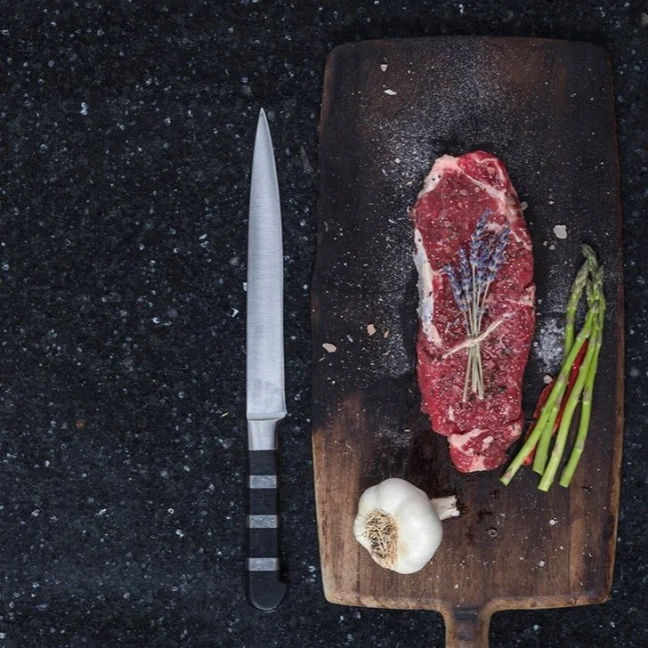 A raw steak seasoned with herbs, a bunch of green asparagus, a garlic bulb, and a large knife on a dark granite countertop, with the meat on a rectangular wooden cutting board.