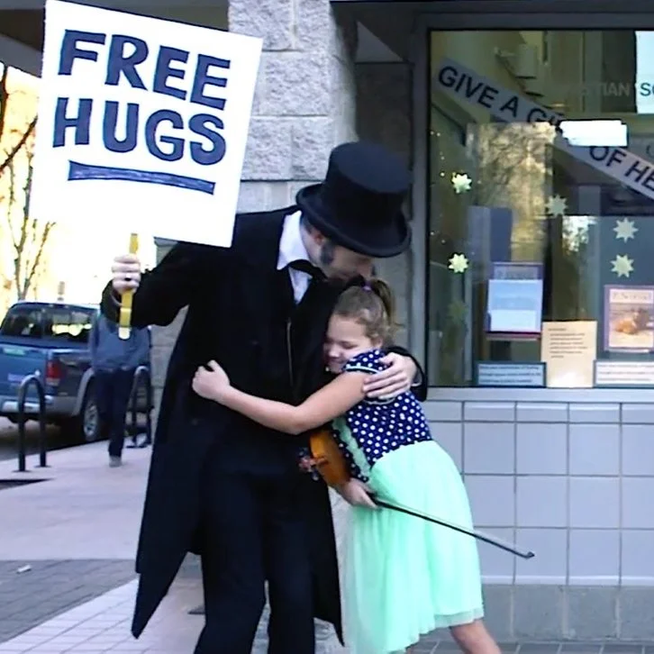 A performer dressed as Charlie Chaplin holding a sign that says "FREE HUGS" and hugging a young girl with brown hair, wearing a blue polka dot top and a light green skirt, outside a building with holiday decorations.