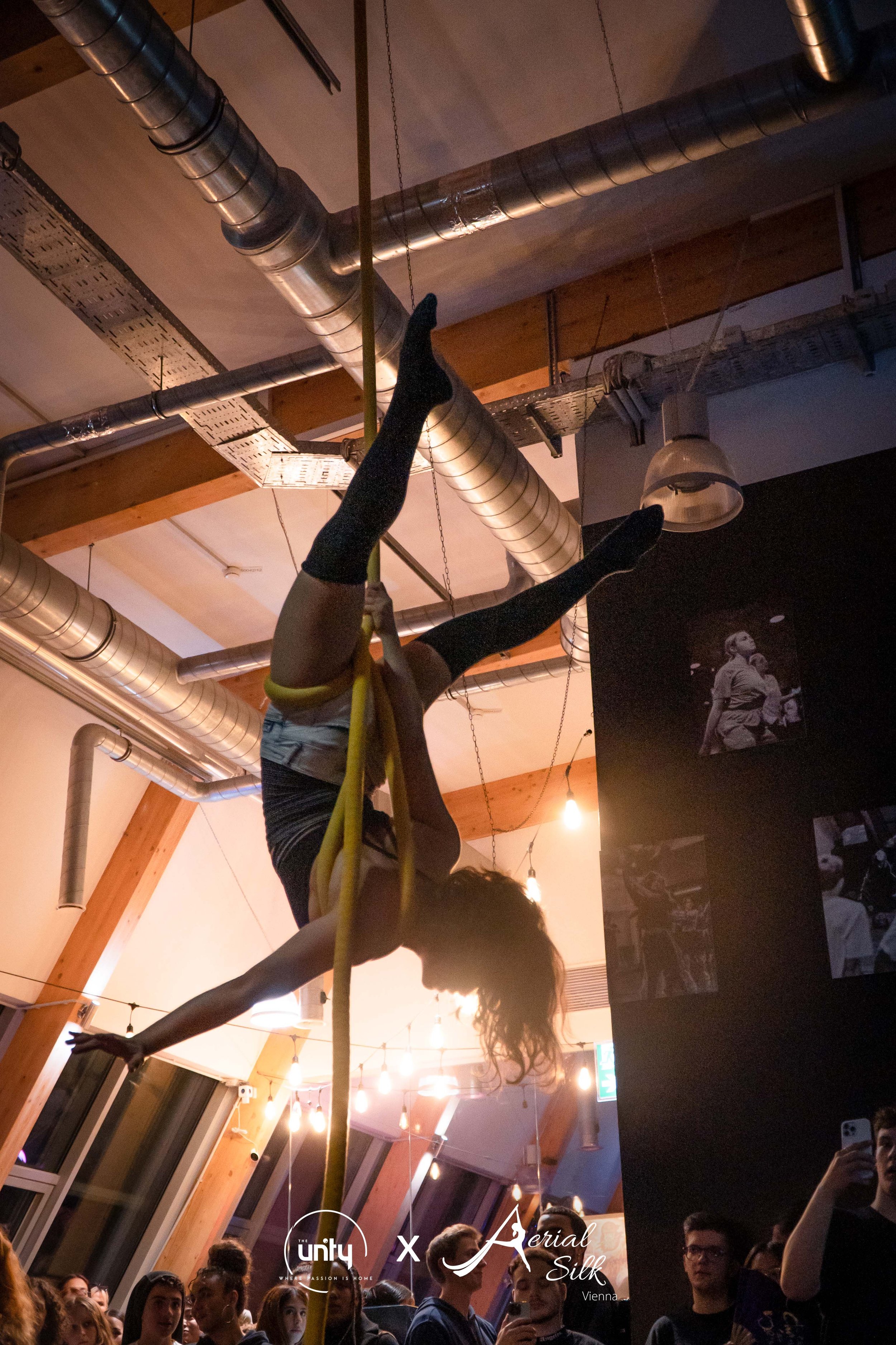 Eine Künstlerin hängt mit schwarzen, langen Socken und kurzen Shorts an einem Seil in der Luft, bei einer Indoor-Event-Veranstaltung mit mehreren Zuschauern.