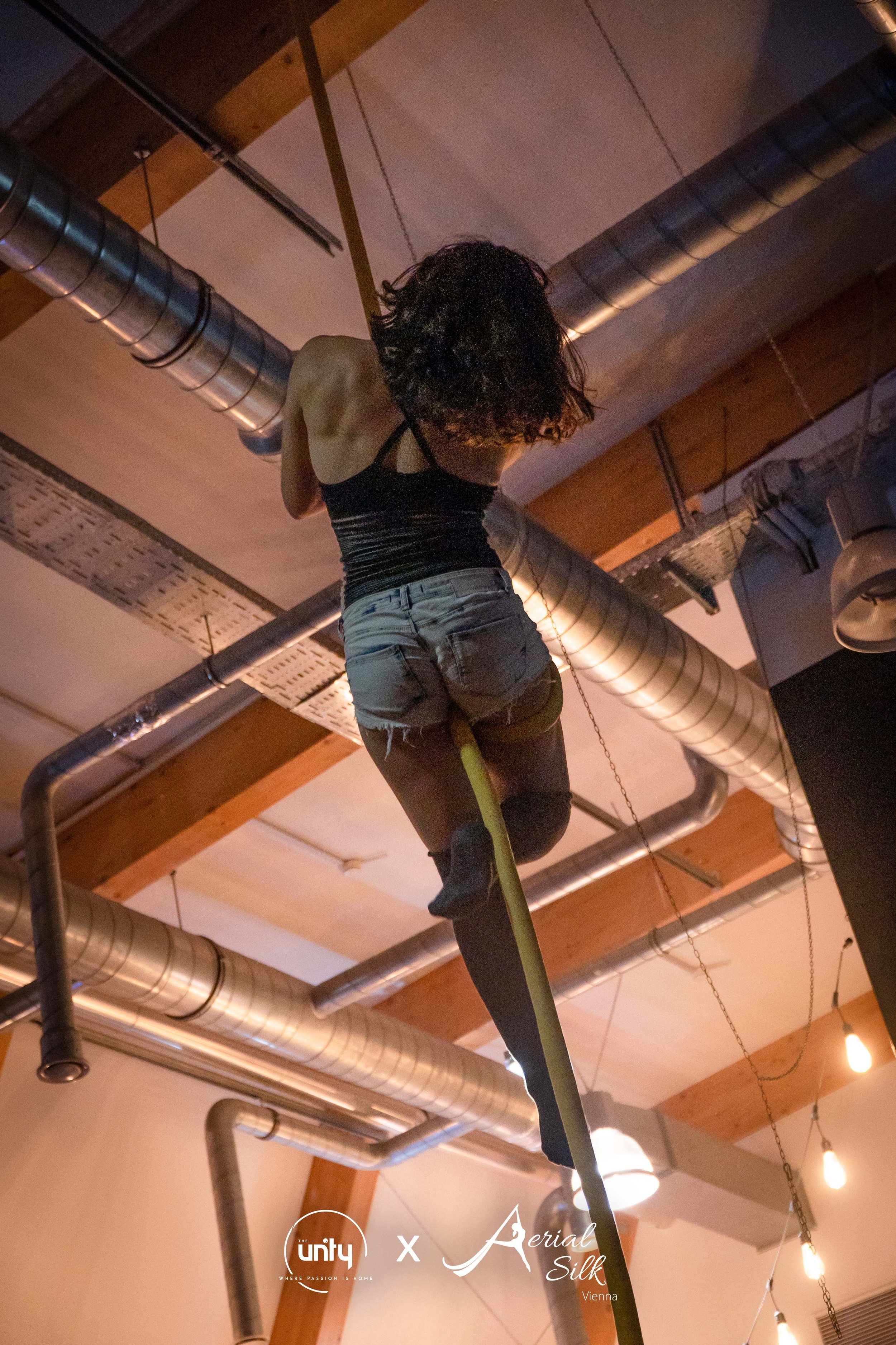 Eine Künstlerin klettert an einem Seil in einer Indoor-Performance, Decken aus Holz und Metallrohre sichtbar, warme Beleuchtung, Logo 'Unity x Aerial Silk Vienna' am unteren Bildrand.