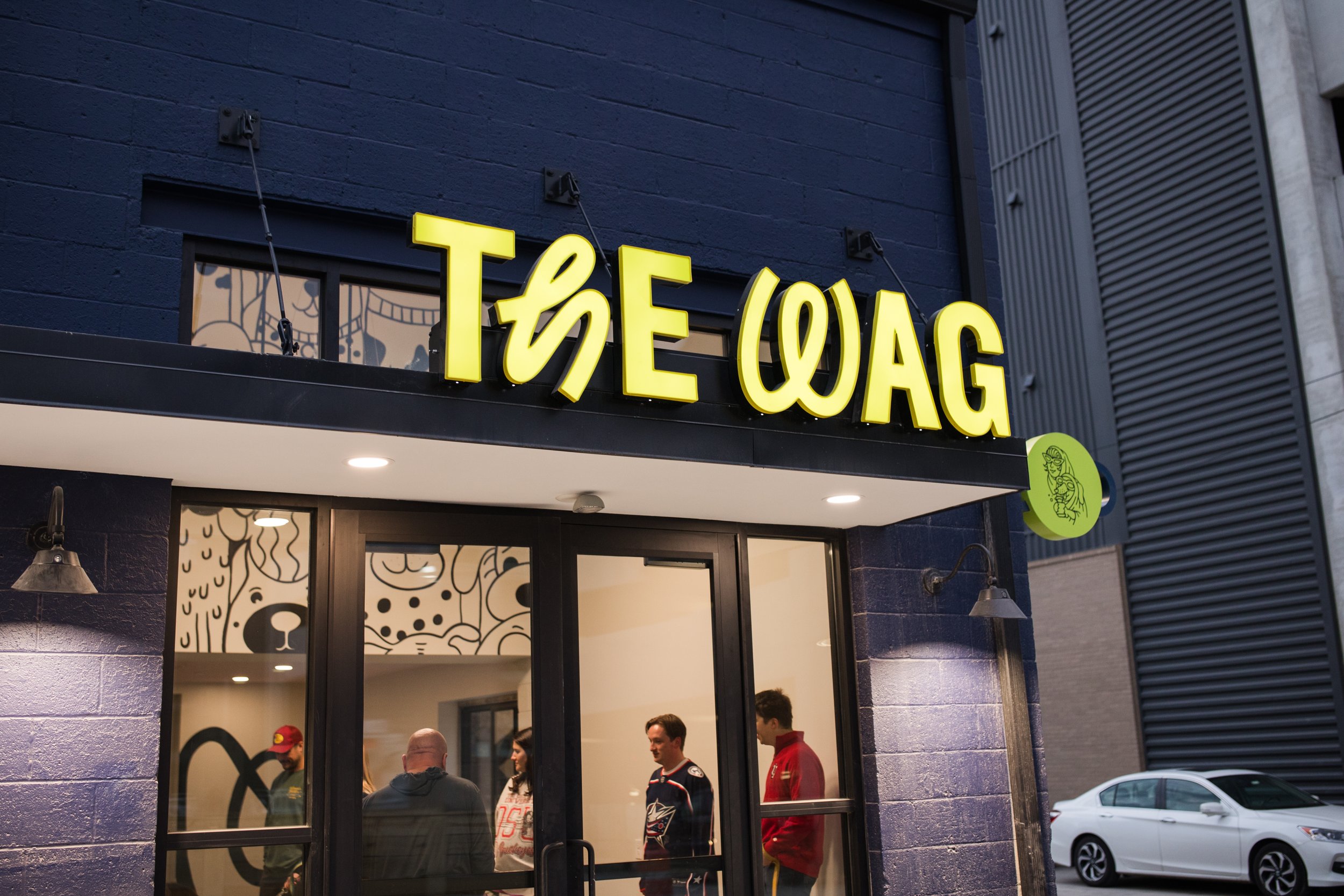 Night view of a storefront with a yellow sign that reads "THE WAG" in stylized letters, and people inside visible through large glass windows, with a mural on the interior wall and a parked white car outside.