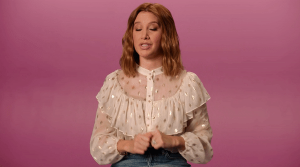 A woman with shoulder-length red hair, wearing a white blouse with gold polka dots, smiling while sitting at a table with a purple background. Next to her is a box and a blister pack of birth control pills.