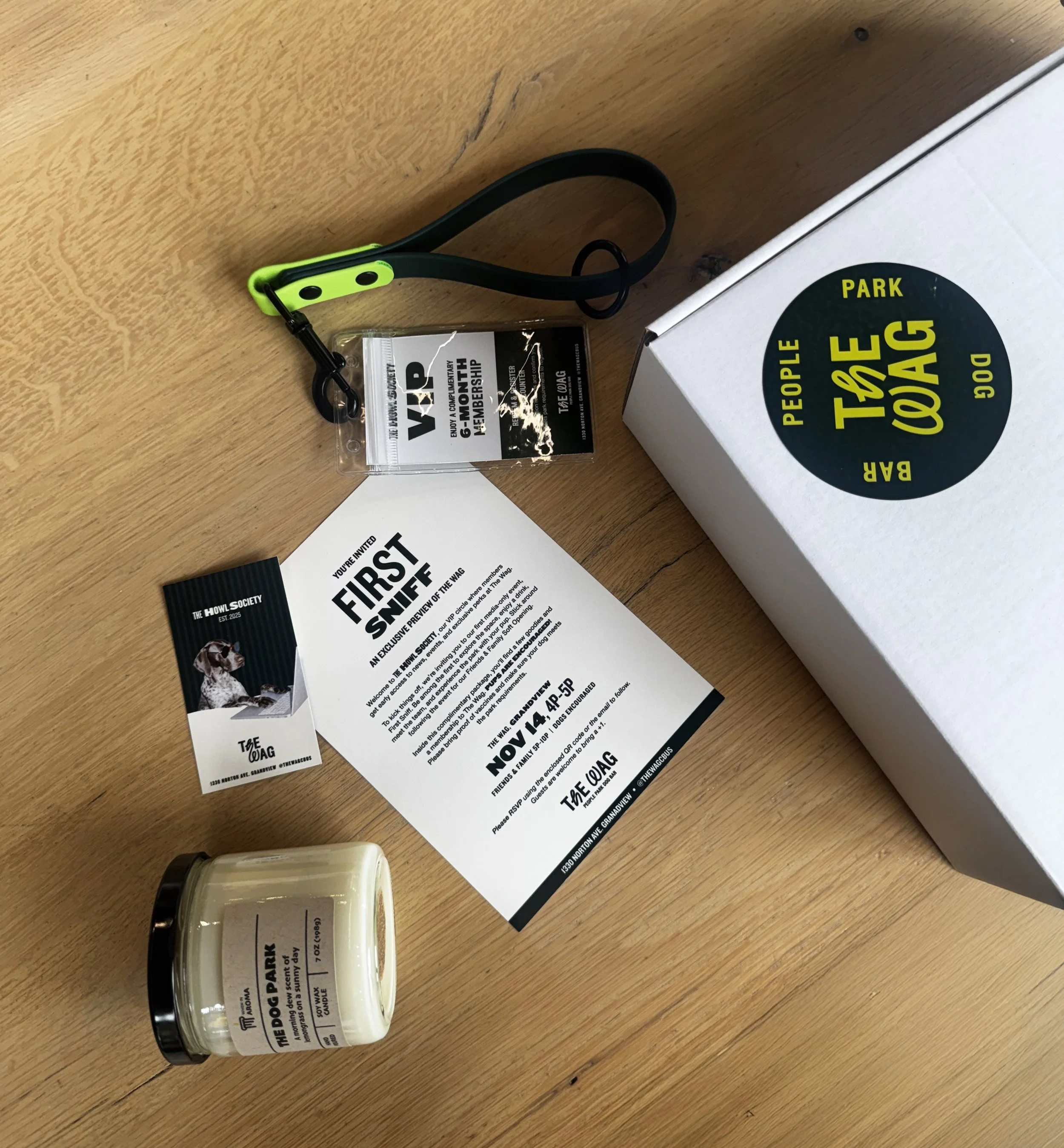 A wooden surface with various items including a green and black lanyard with a badge, a packet for a dog membership, a flyer for the Dog Park, a candle, a small real estate flyer, and a large white box with a black and yellow circular sticker that says "People Bark Park".