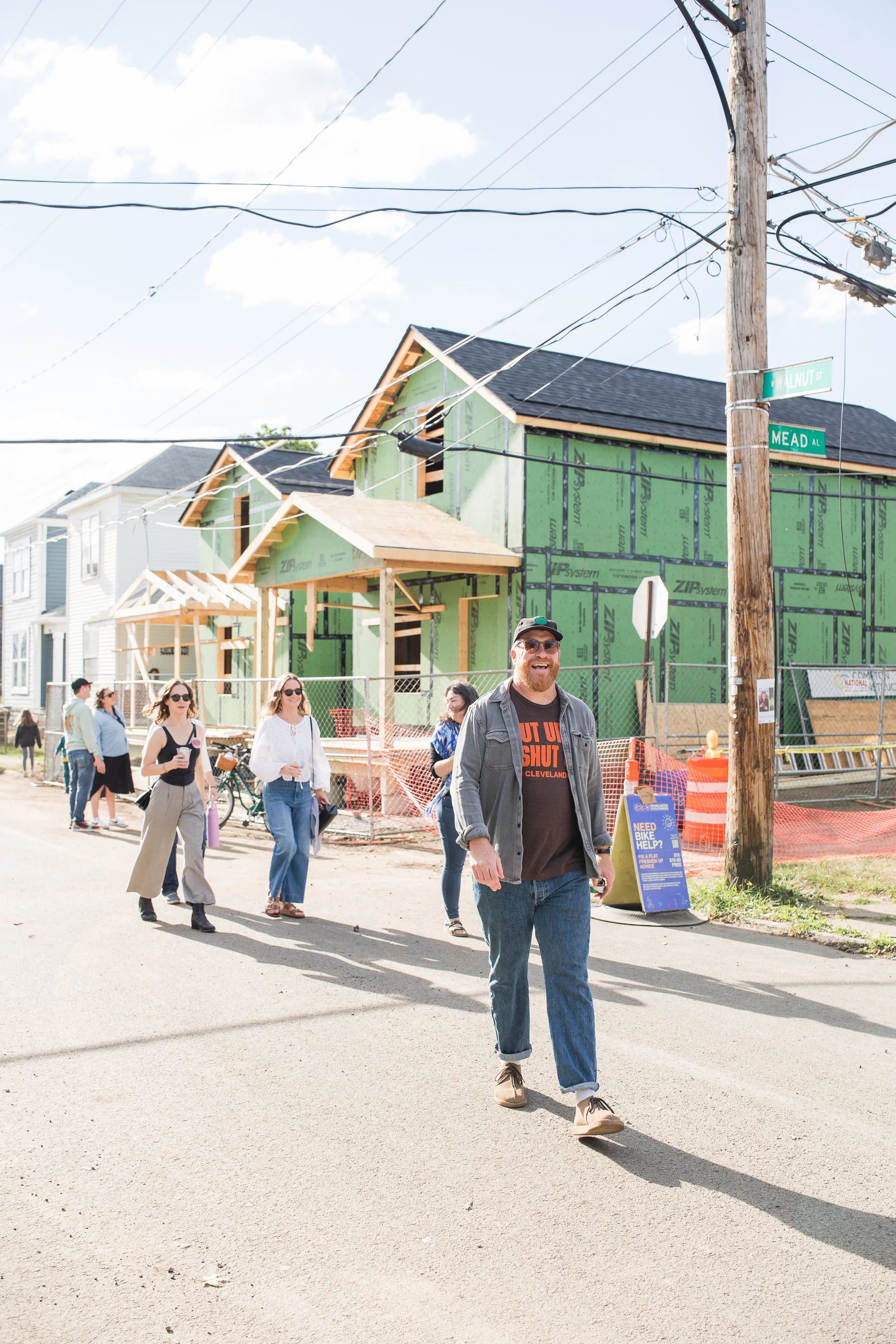 People walking on a street past a construction site with new houses being built, and a man in the front smiling and wearing sunglasses.