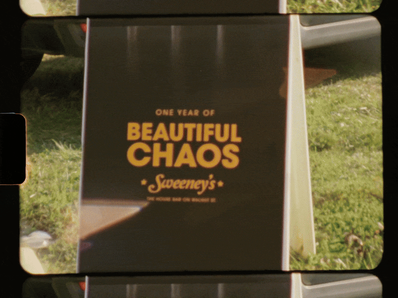 Signboard celebrating one year of Sweeney's with the phrase "Beautiful Chaos" in large yellow letters, placed outdoors on a grassy area.