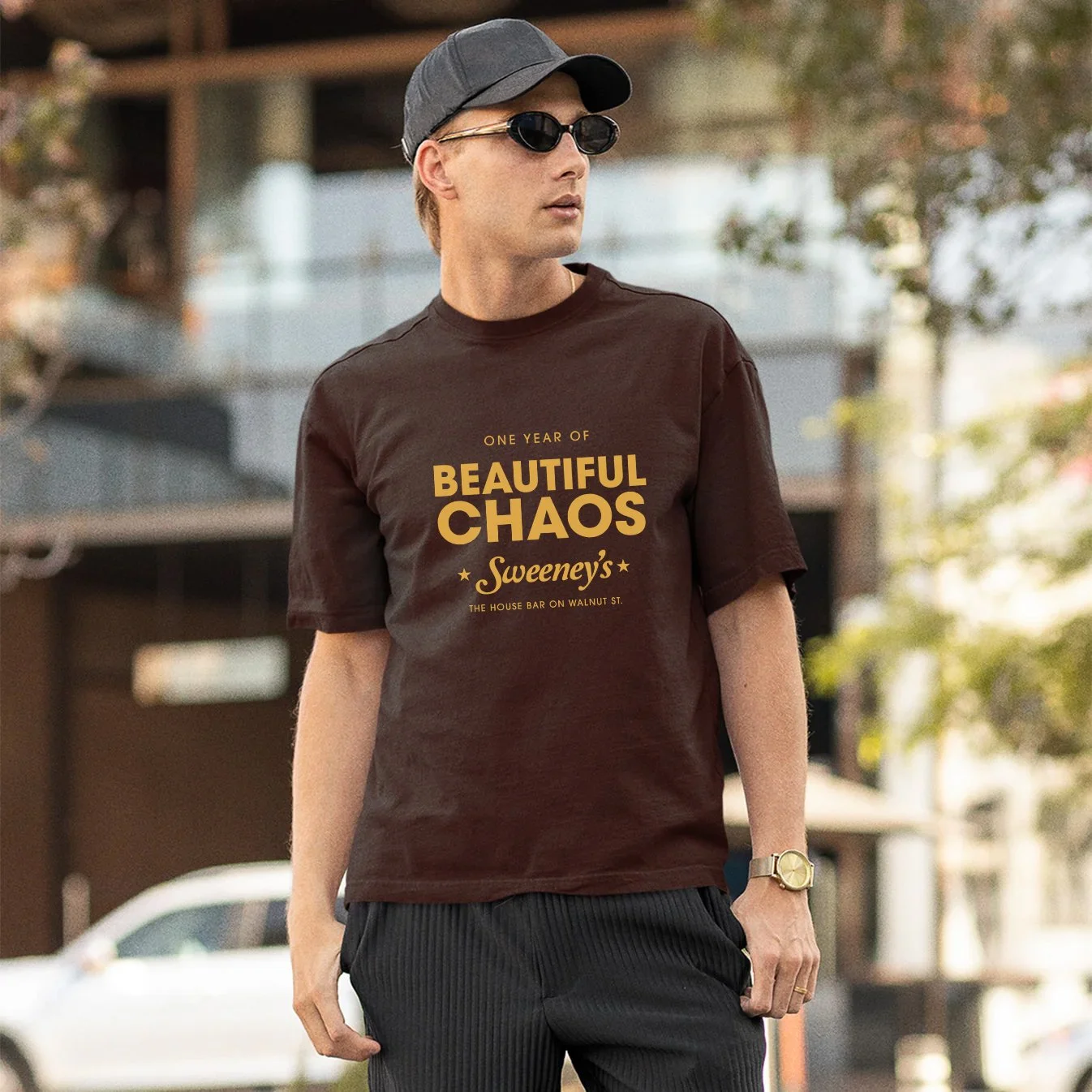 A man wearing a black cap, dark sunglasses, a brown T-shirt with yellow text, black pants, and a wristwatch, standing outdoors in front of trees and buildings.