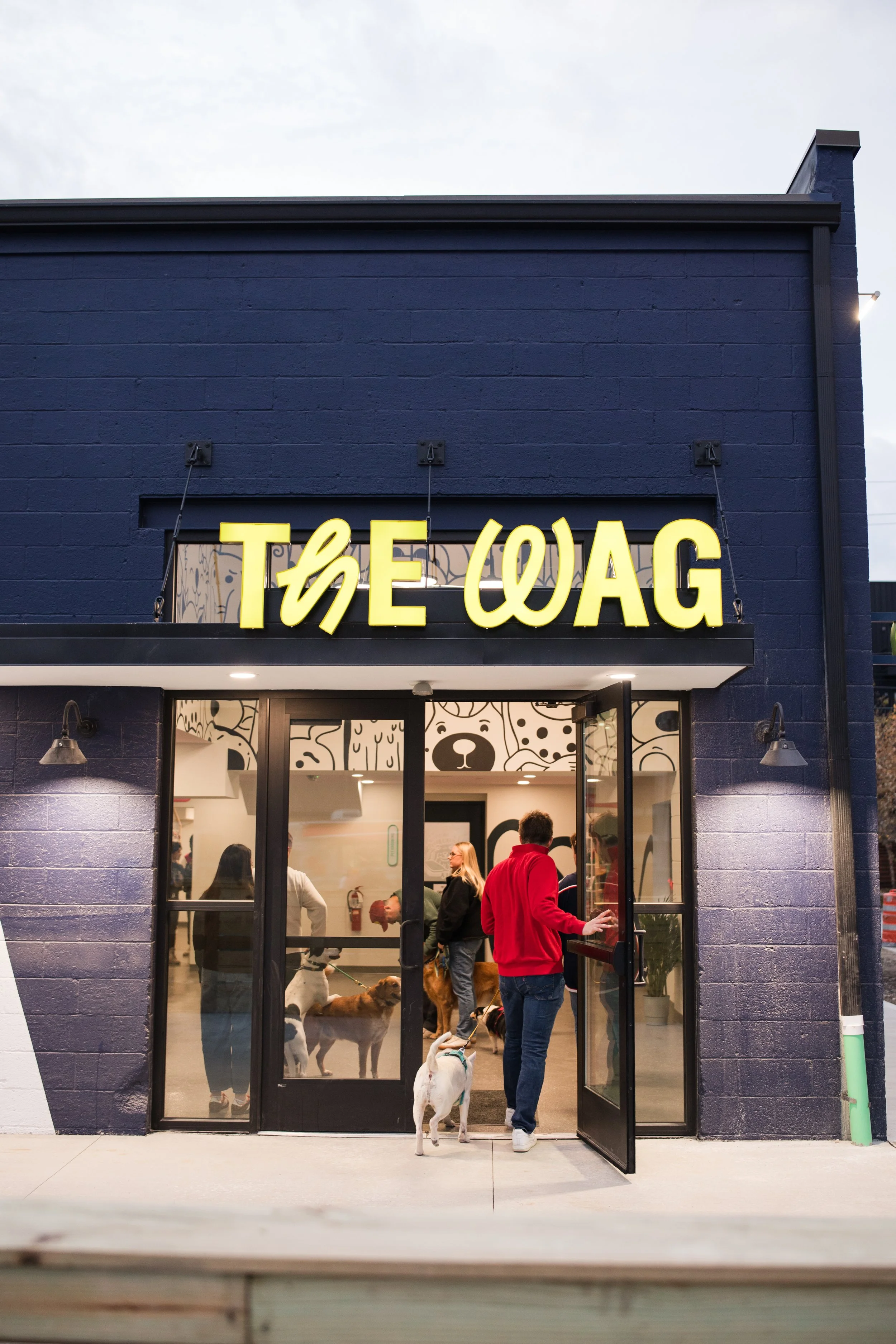 People and dogs entering The WAG pet store, with a dark exterior and large glass doors.