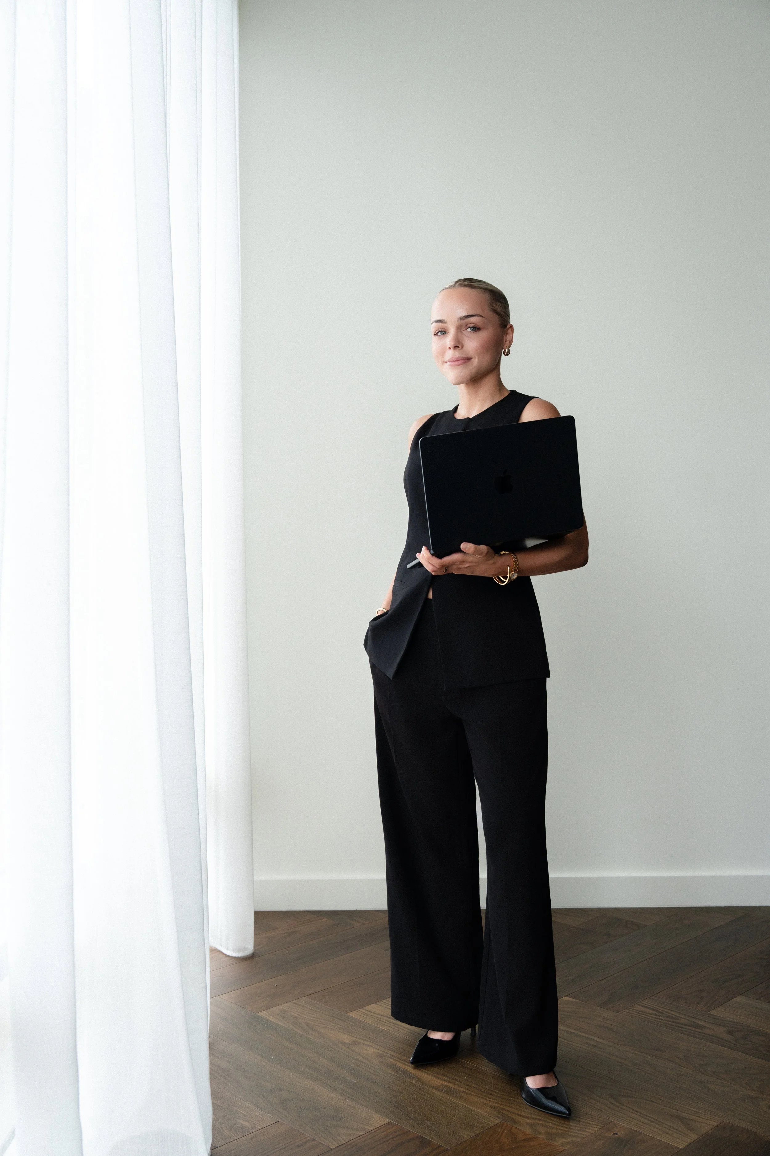 A woman in a black sleeveless top and wide-legged black pants standing indoors, holding a black laptop with a silver Apple logo, and looking confidently at the camera. White curtains are visible on the left, and the room has a wooden floor and light gray wall.