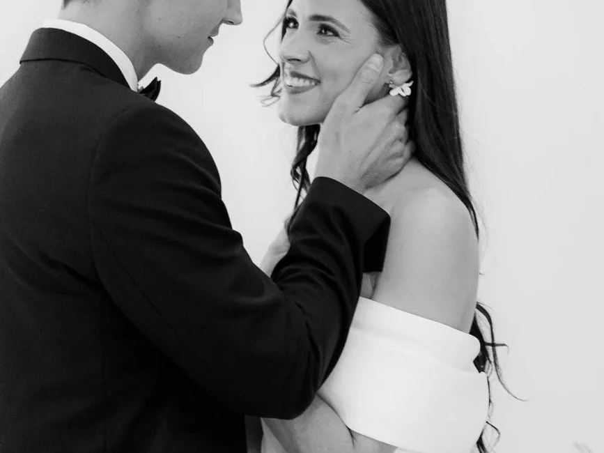 Ein Mann in einem schwarzen Anzug hält eine Frau in einem weißen Kleid liebevoll im Arm und schaut sie zärtlich an, beide lächeln.