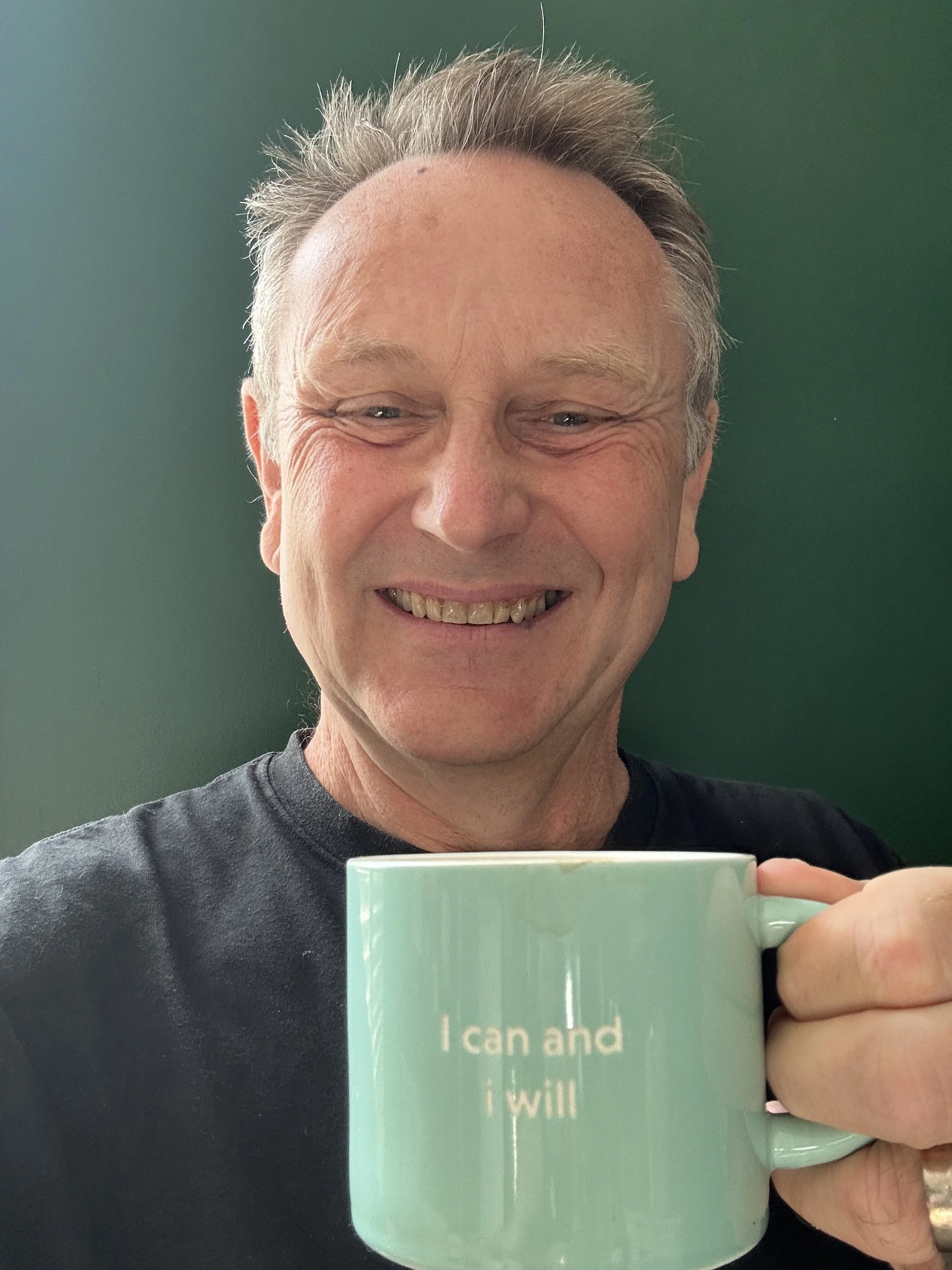 A smiling person taking a selfie in front of a green background while holding a turquoise mug with white text.