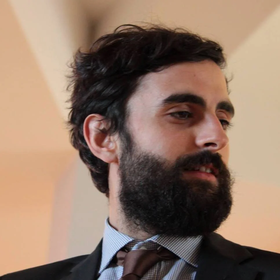 Tommaso Scavone, uomo con barba scura, capelli mossi e abbigliamento formale, che indossa una giacca e una cravatta, in un ambiente interno.