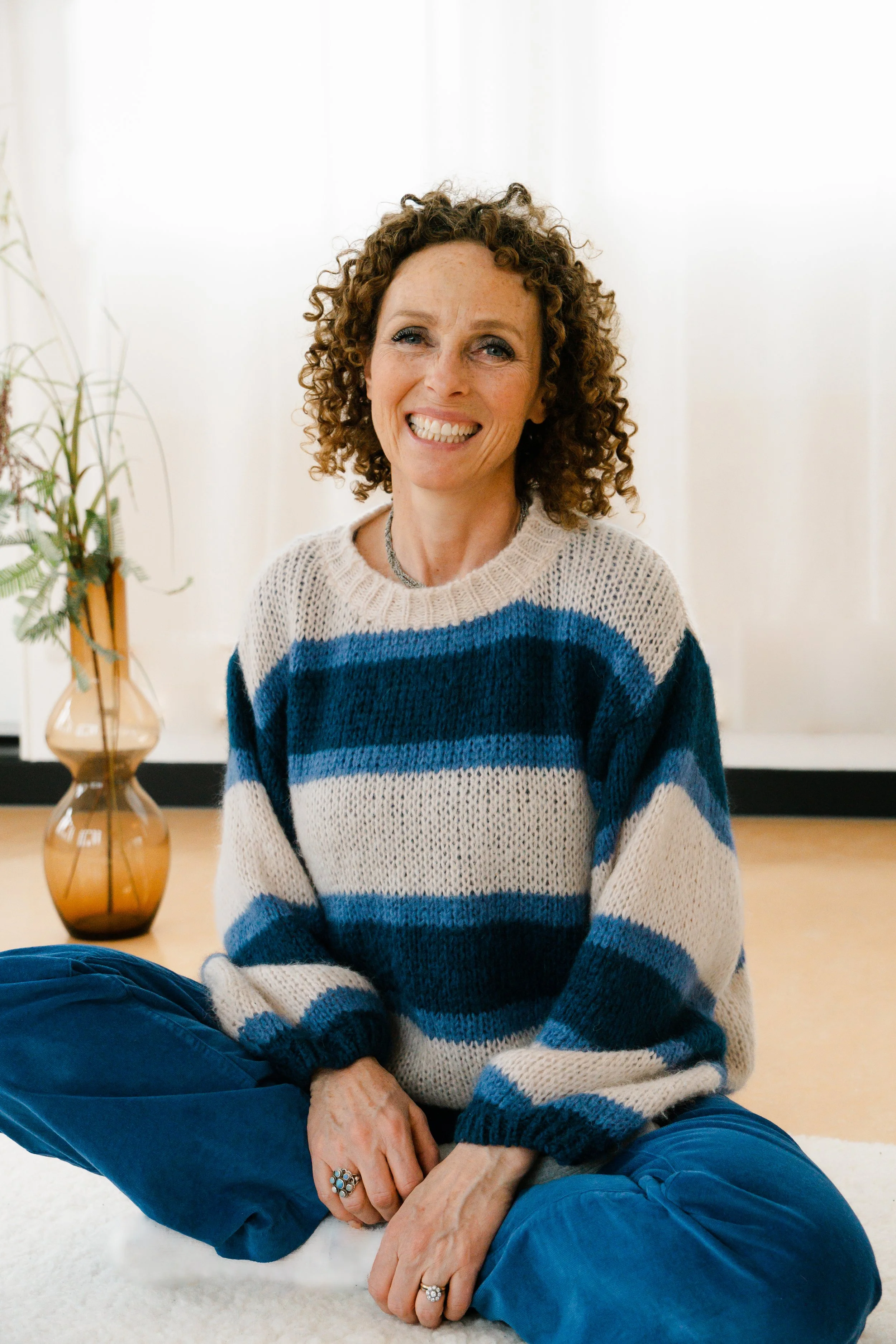 Een glimlachende vrouw met krullend haar zit gekruist op de vloer, gekleed in een gebreide trui met blauwe en beige strepen, met een plant in een vaas op de achtergrond.
