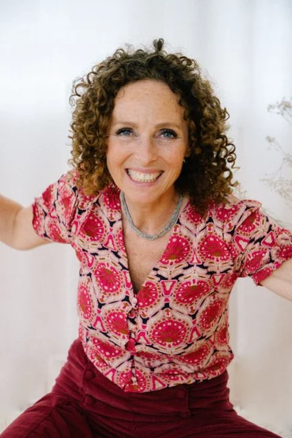 Vrolijke vrouw met gekrulde haren die lacht en zich uitstrekt, zittend voor een lichte achtergrond.