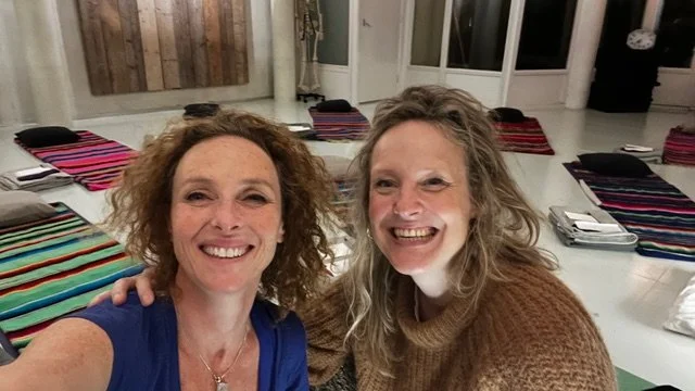 Twee vrouwen die in een kamer poseren voor een selfie, met kleurrijke gestreepte zitzakken en stapels papieren op de achtergrond.