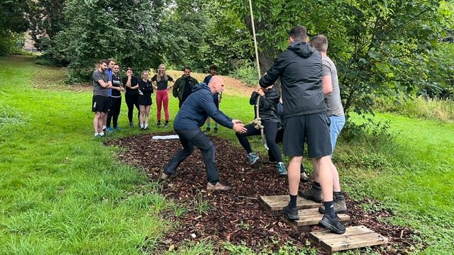 Groep mensen die deelnemen aan een teamactiviteit buiten, waarbij ze een touw trekken en over een houten brug lopen, omringd door groen bomen en gras.