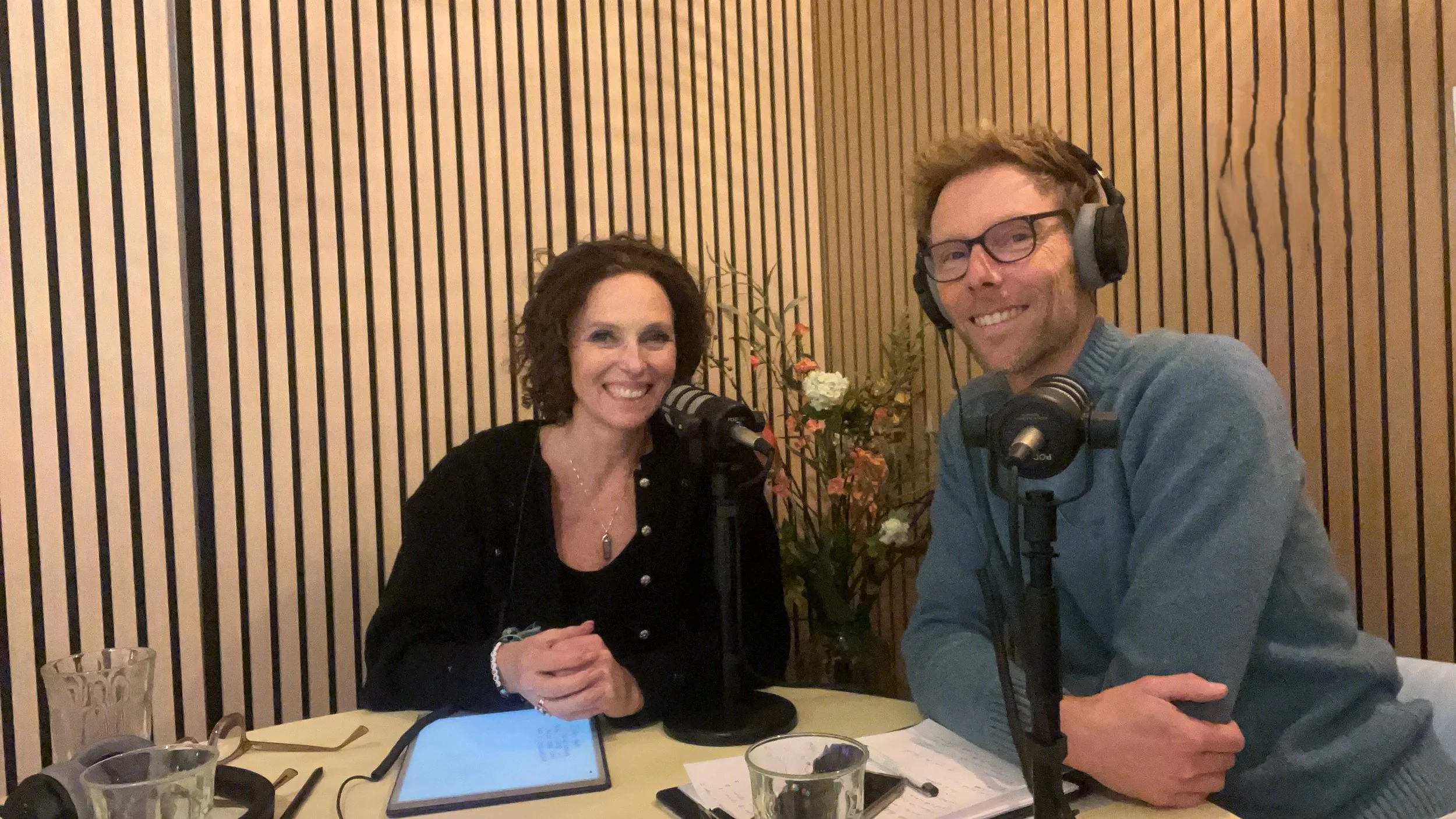 Twee mensen zitten aan een tafel met microfoons, koptelefoons en notities, lachend, in een geluidsstudio met houten wandpanelen.
