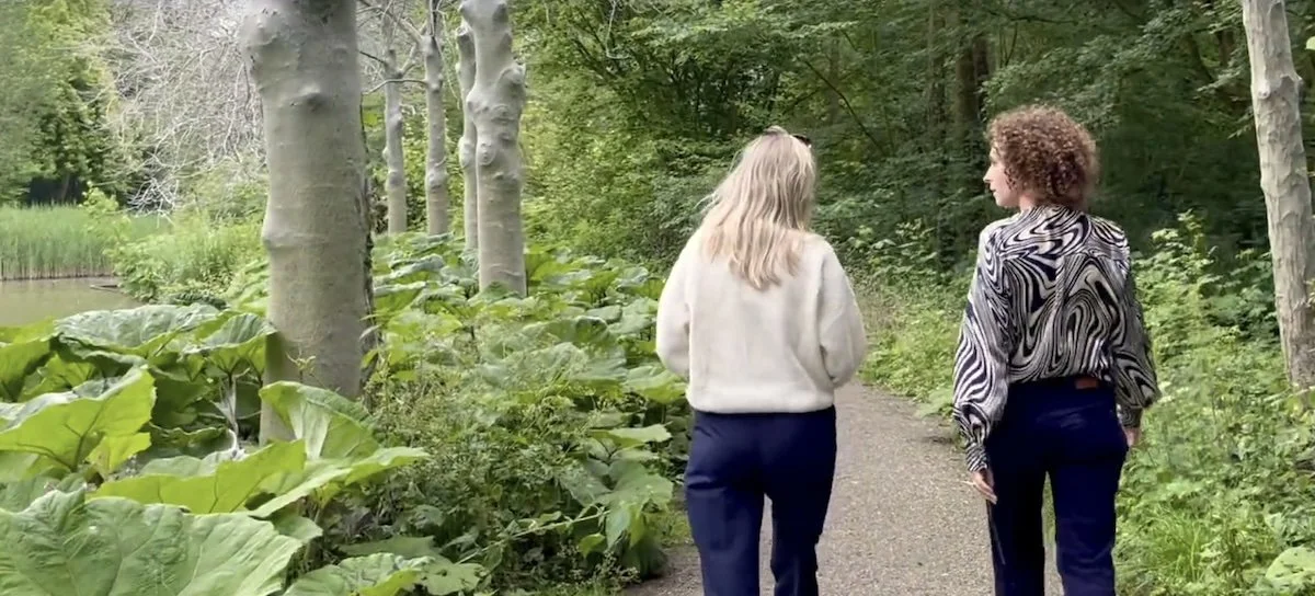 Twee vrouwen wandelen op een bospad, omringd door groene planten en bomen.