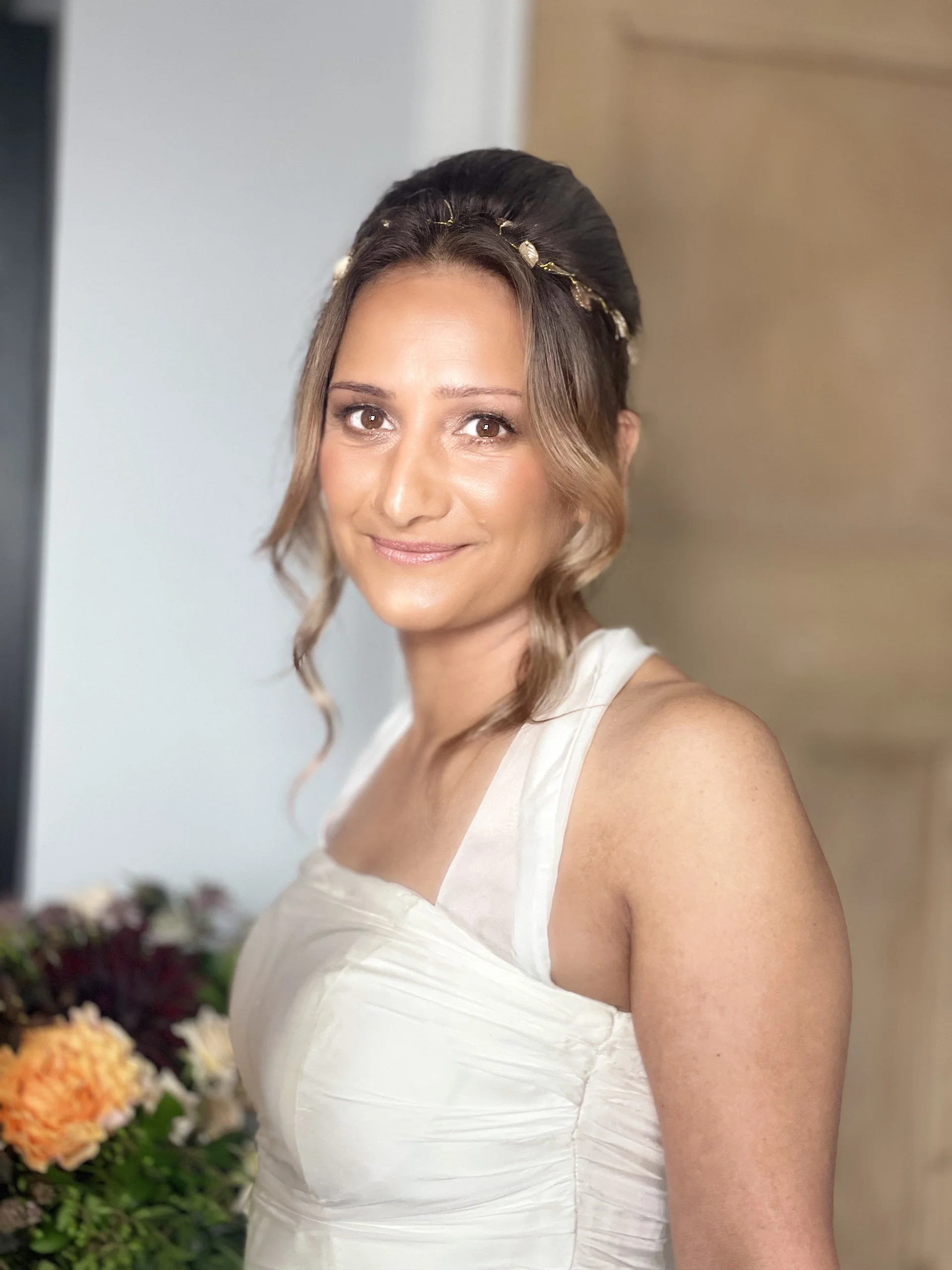A woman with wavy hair styled up with a gold hairband, wearing a white dress, smiling, standing in front of a floral arrangement.