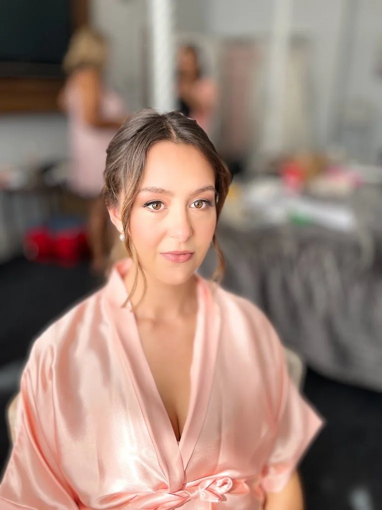 A young woman with brown hair and brown eyes, wearing a pink satin robe, sitting indoors with a blurred background of two women in a kitchen setting.