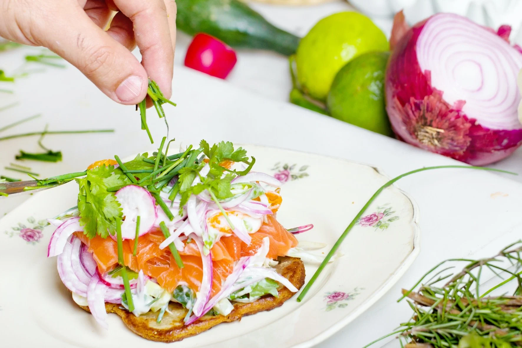 Hand garnishing open-faced sandwich with herbs, smoked salmon, and vegetables, surrounded by onions, limes, and a plate.
