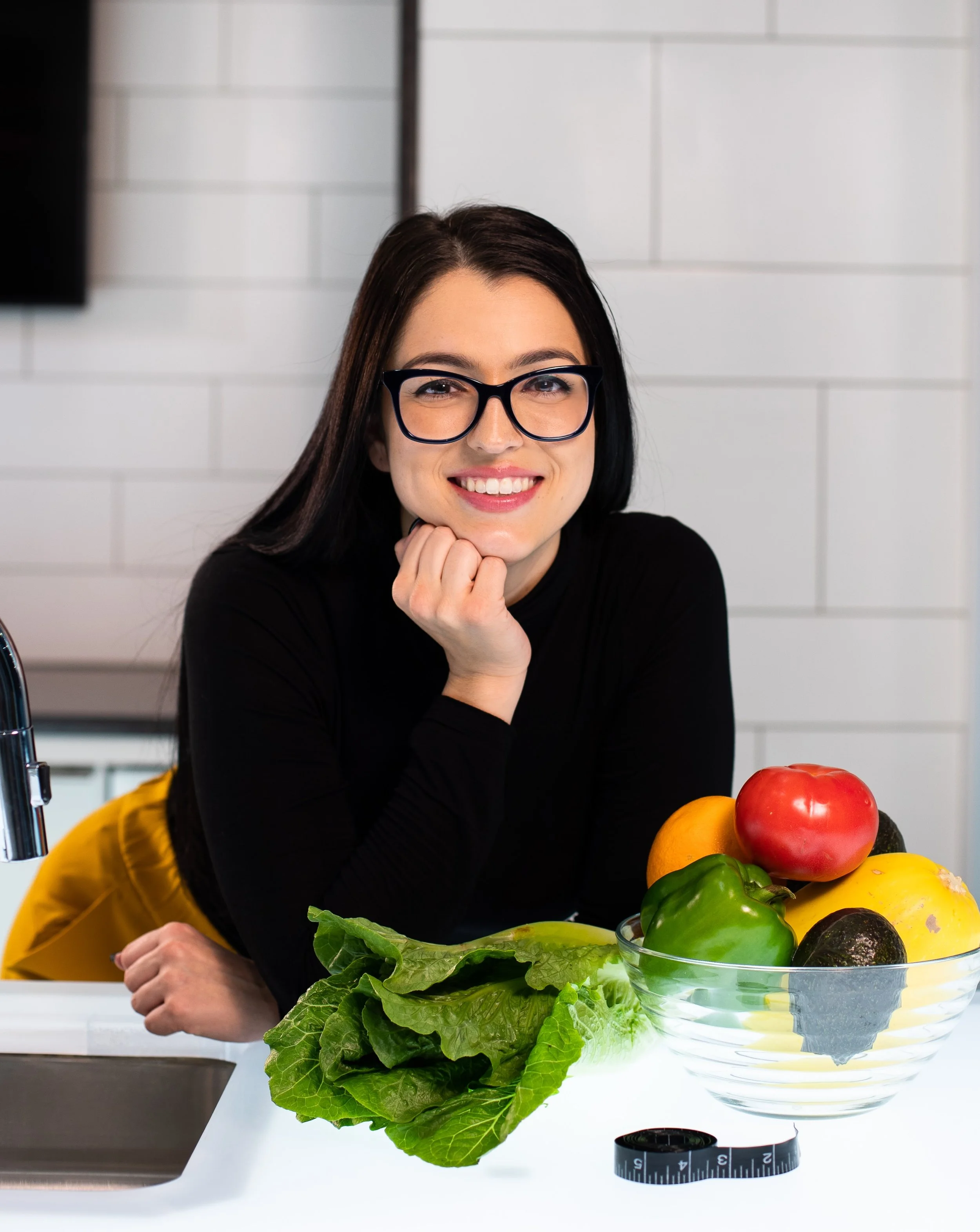 Smiling person with glasses next to a bowl of fresh vegetables including tomato, bell pepper, avocado in a modern kitchen.