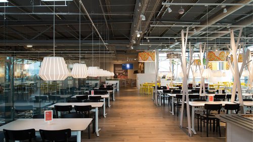 Inside a modern, empty restaurant with white pendant lights, black chairs, white tables, and yellow chairs in the background.