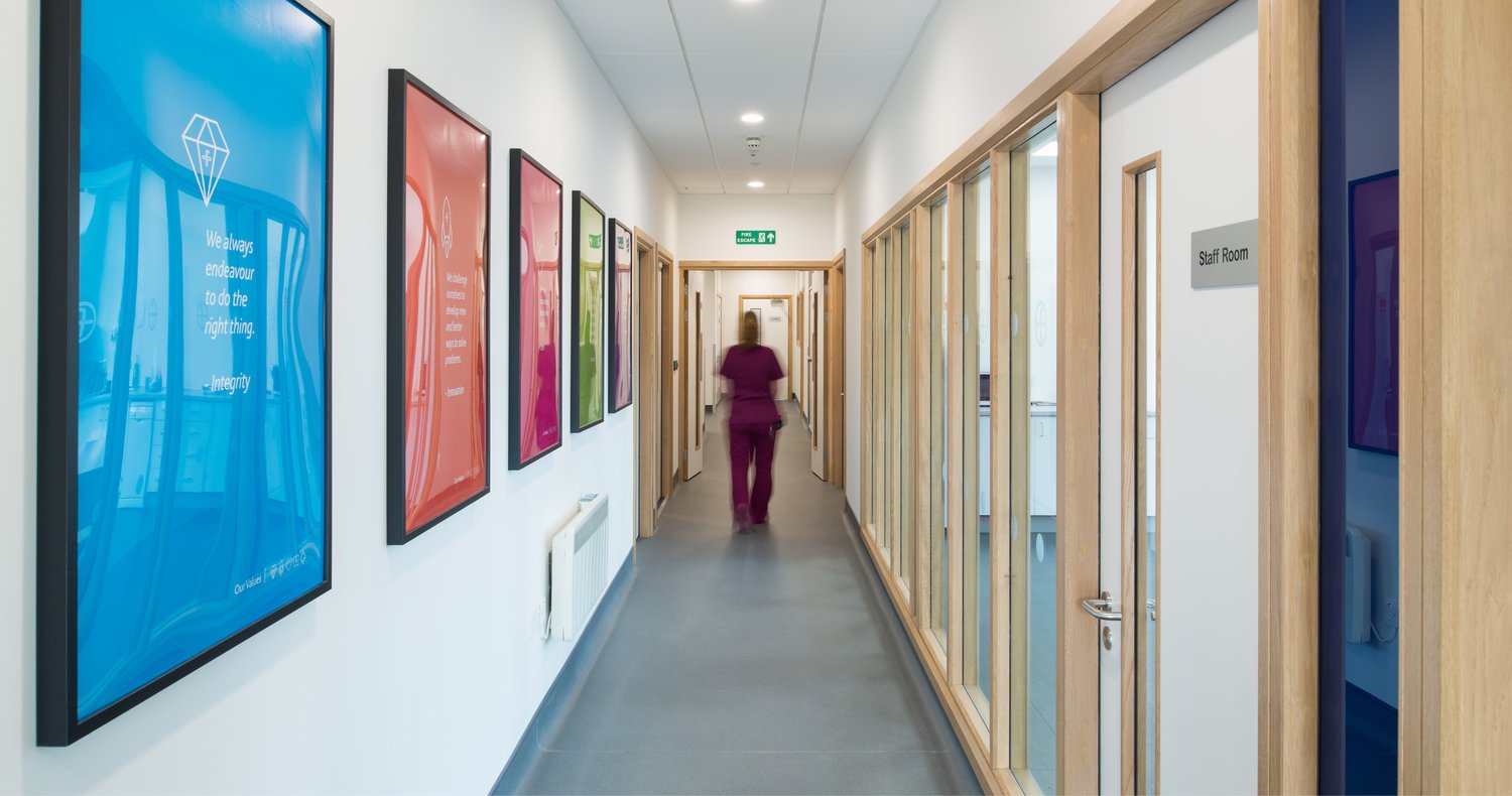 Hospital corridor with colorful framed posters on the left wall, doorways on the right with glass panels, and a nurse walking towards the exit sign at the end of the hallway.