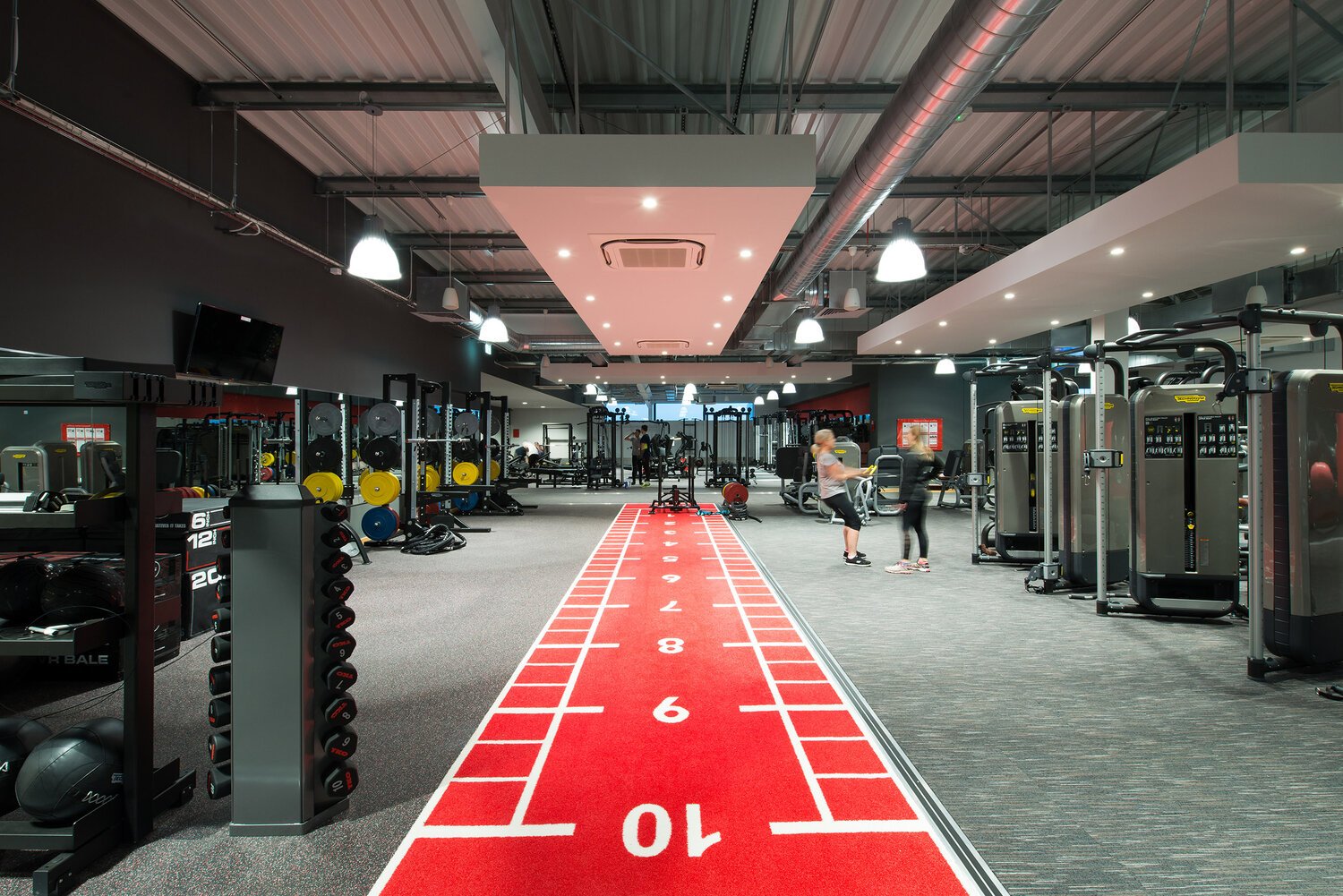 Indoor gym with workout equipment, weight racks, cardio machines, and a red running track in the center. People are exercising and socializing in the background.