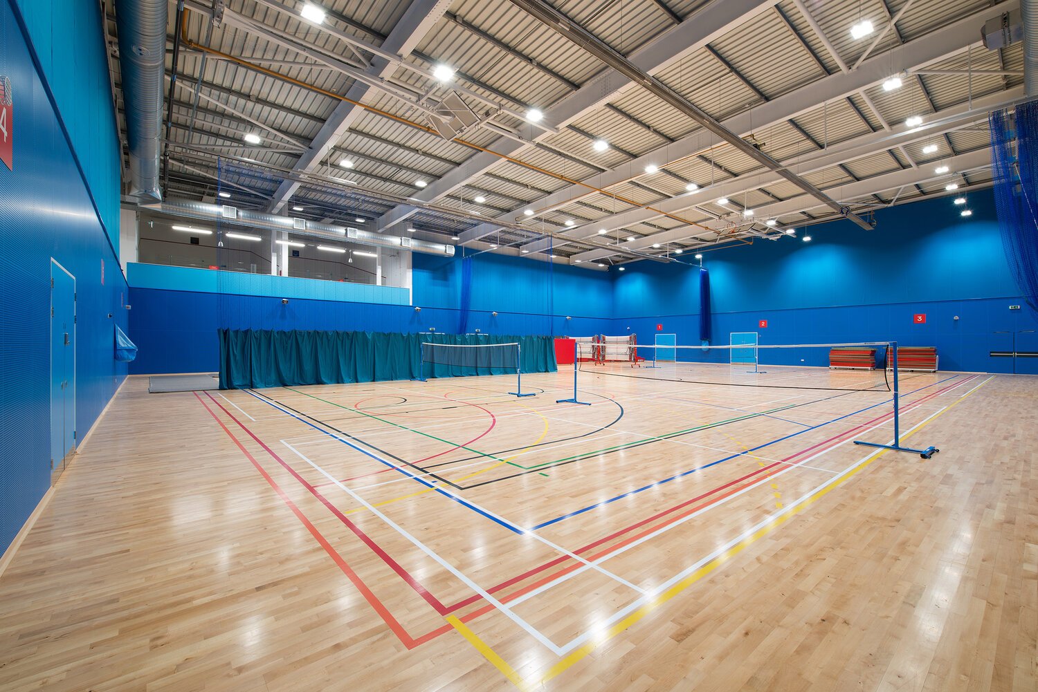 Empty indoor sports court with badminton nets, wooden flooring, blue walls, and overhead lighting.