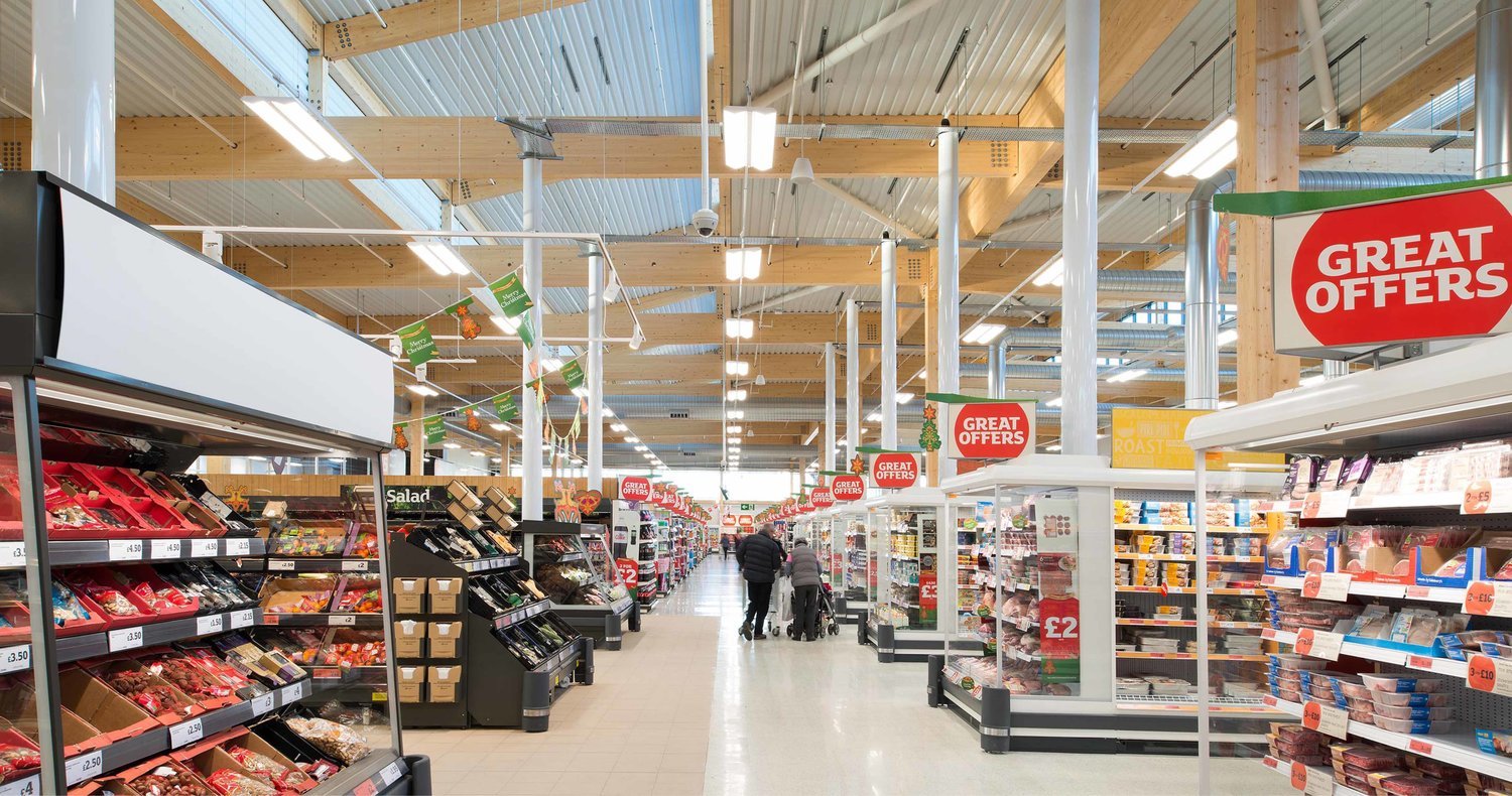 Inside a grocery store with shelves stocked with meats and other products, signs advertising great offers, and shoppers including two adults and a child in the distance.