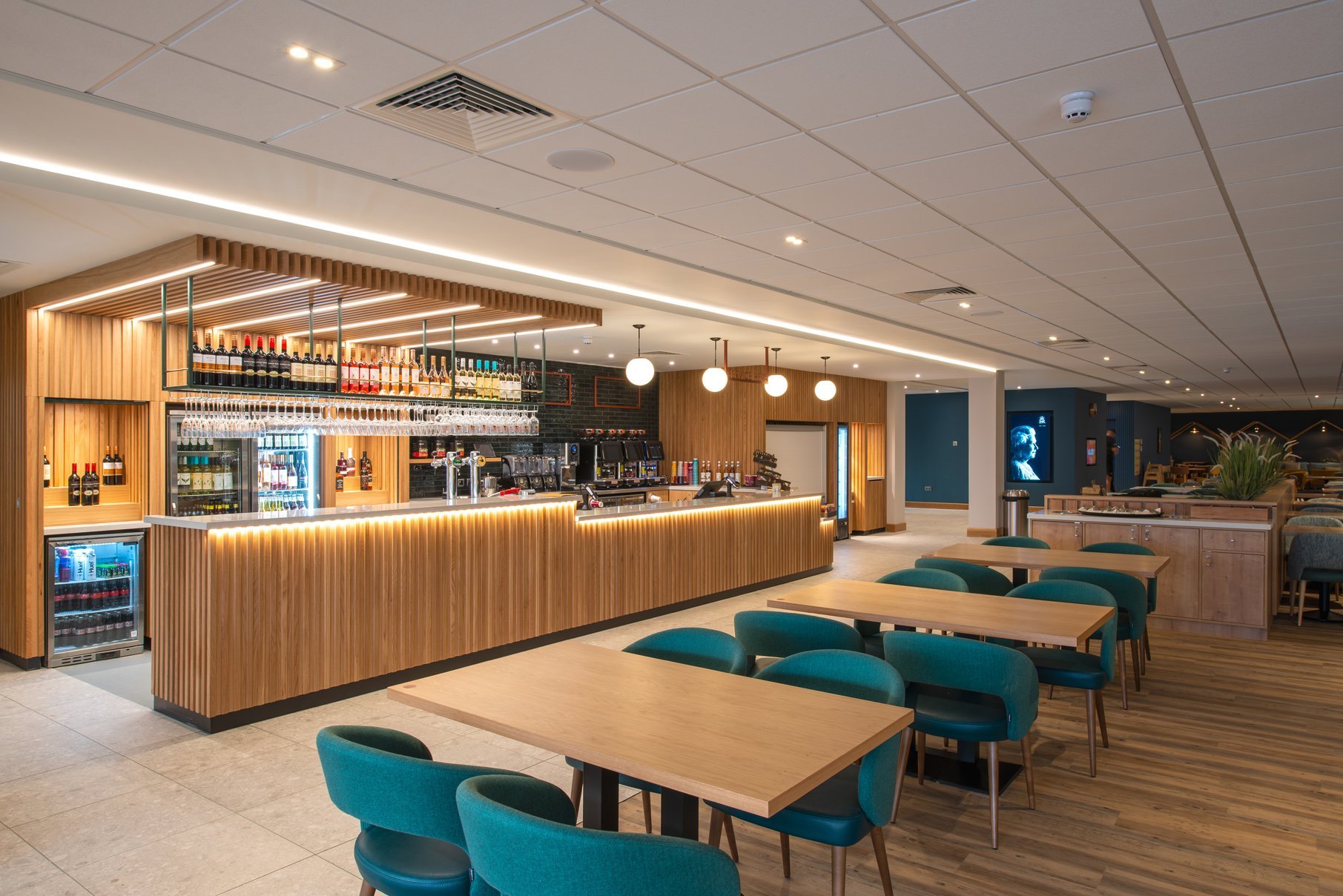 Empty restaurant with a wooden bar, teal chairs, and a lounge area, featuring modern lighting and decor.